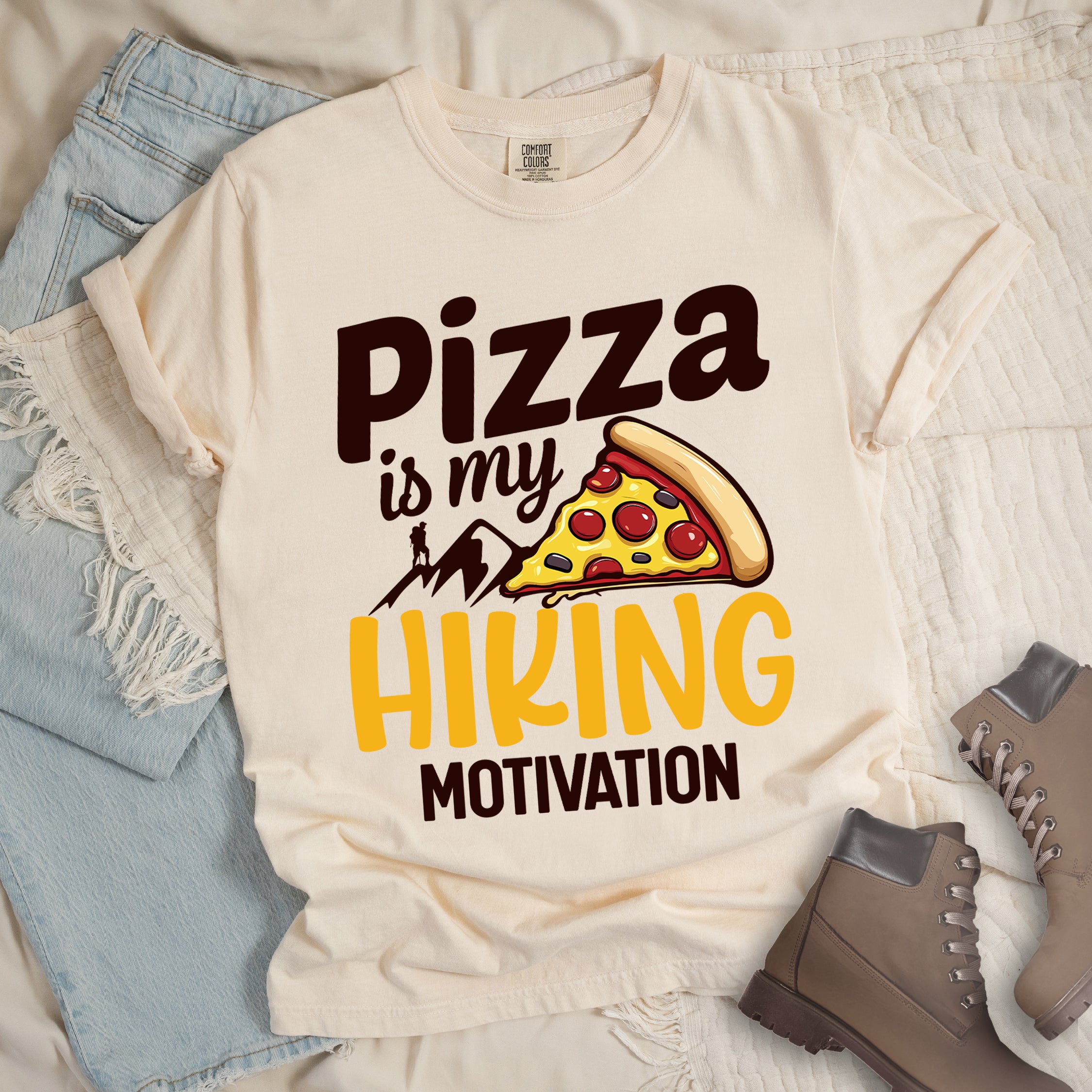 Ivory (Cream White) t-shirt with deep brown and yellow text reading "Pizza is my HIKING MOTIVATION". "Pizza" appears in large deep brown letters at the top, "is my" in smaller deep brown script, "HIKING" in yellow at the bottom, and "MOTIVATION" in deep brown. Features a colorful pizza slice with pepperoni toppings, deep brown mountain silhouettes with a small hiker figure on a peak. The design humorously connects pizza as the incentive for hiking activities.
