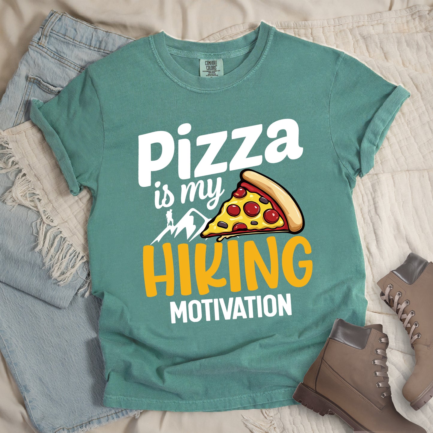 Light Green t-shirt with white and yellow text reading "Pizza is my HIKING MOTIVATION". "Pizza" appears in large white letters at the top, "is my" in smaller white script, "HIKING" in yellow at the bottom, and "MOTIVATION" in white. Features a colorful pizza slice with pepperoni toppings, white mountain silhouettes with a small hiker figure on a peak. The design humorously connects pizza as the incentive for hiking activities.