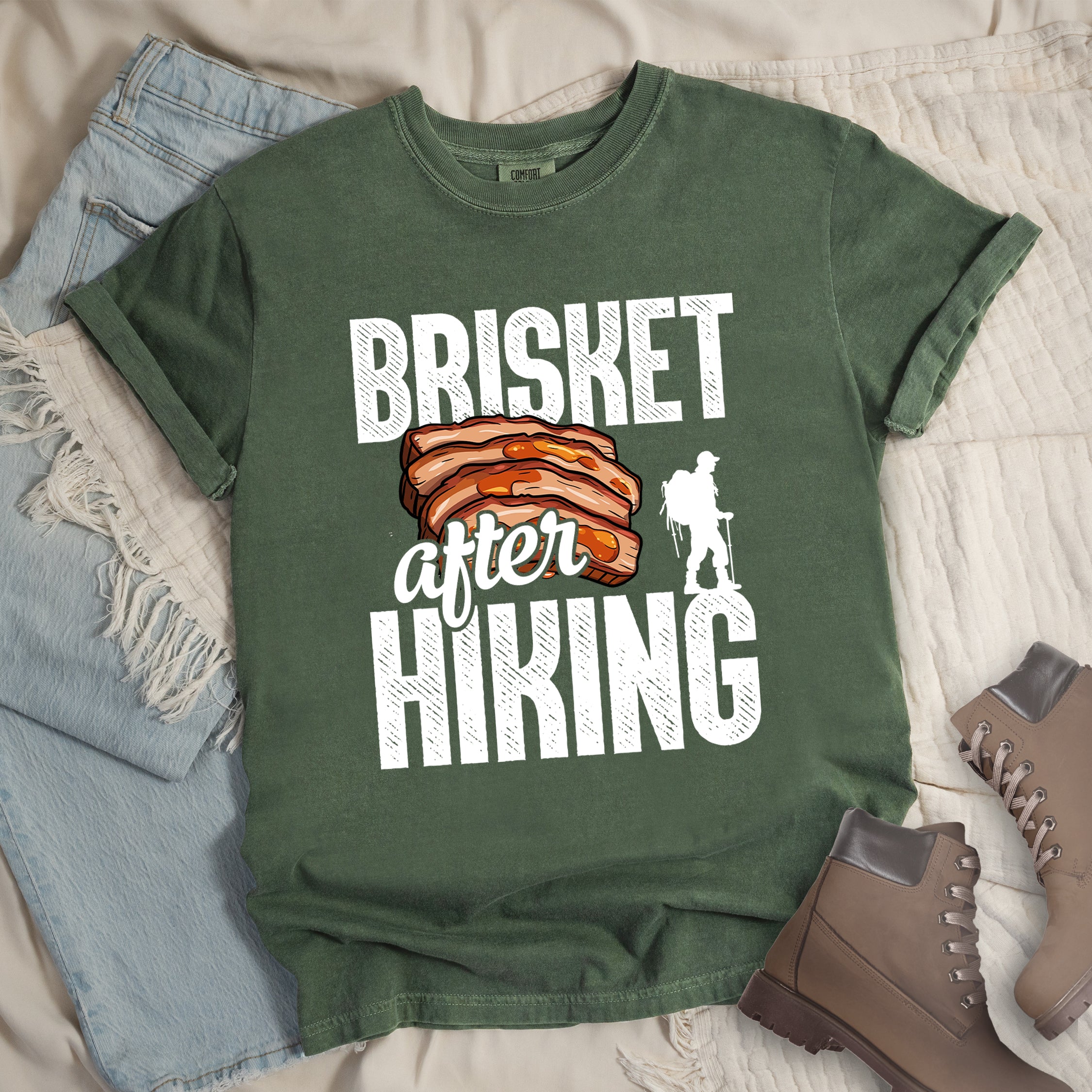 Moss (Dark Green) t-shirt with white distressed text reading "BRISKET after HIKING". "BRISKET" appears in large white letters at the top, "after" in white script in the middle, and "HIKING" in large white letters at the bottom. Features a detailed illustration of sliced brisket meat in orange-brown tones in the center and a white silhouette of a hiker with walking poles on the right side.