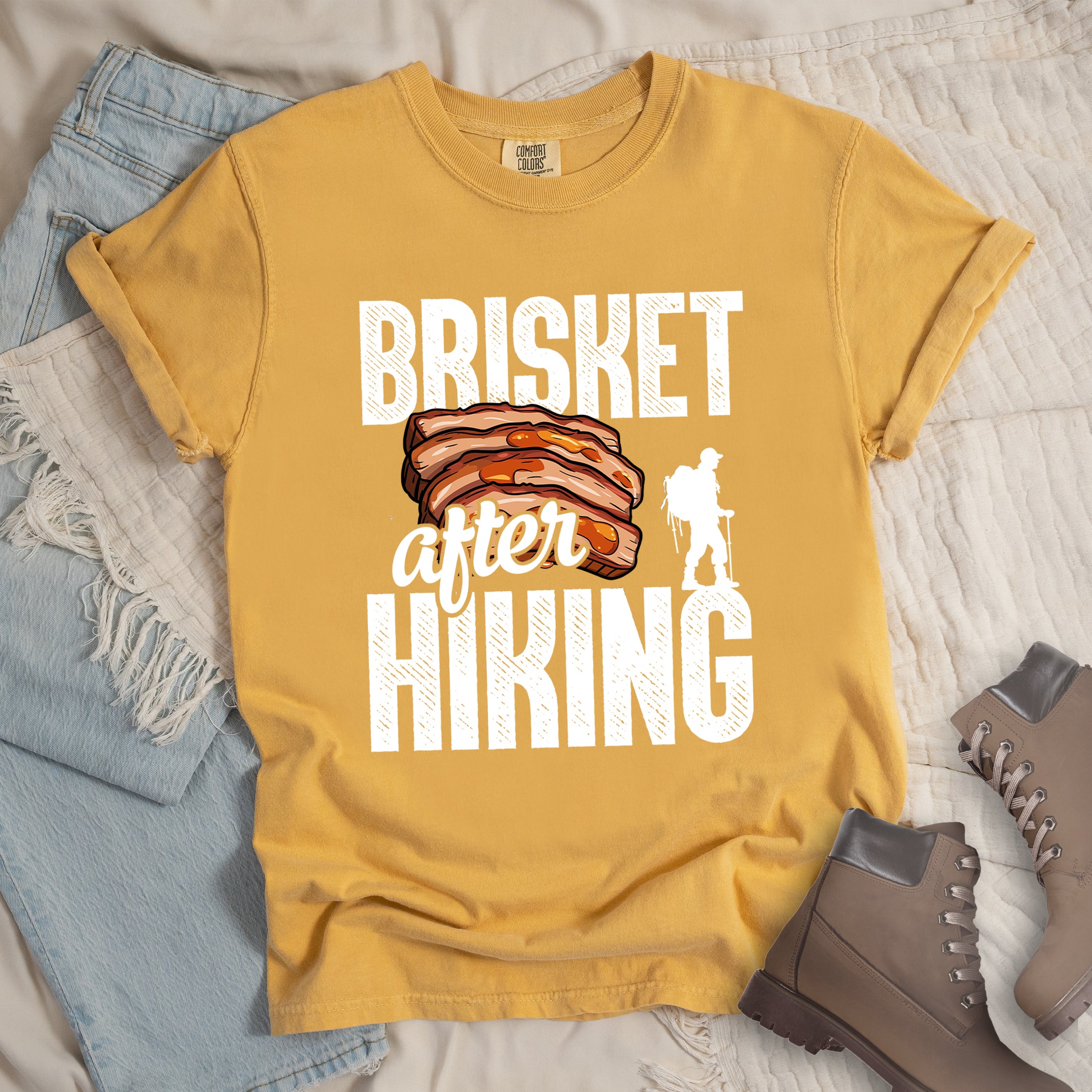 Mustard (Yellow) t-shirt with white distressed text reading "BRISKET after HIKING". "BRISKET" appears in large white letters at the top, "after" in white script in the middle, and "HIKING" in large white letters at the bottom. Features a detailed illustration of sliced brisket meat in orange-brown tones in the center and a white silhouette of a hiker with walking poles on the right side.