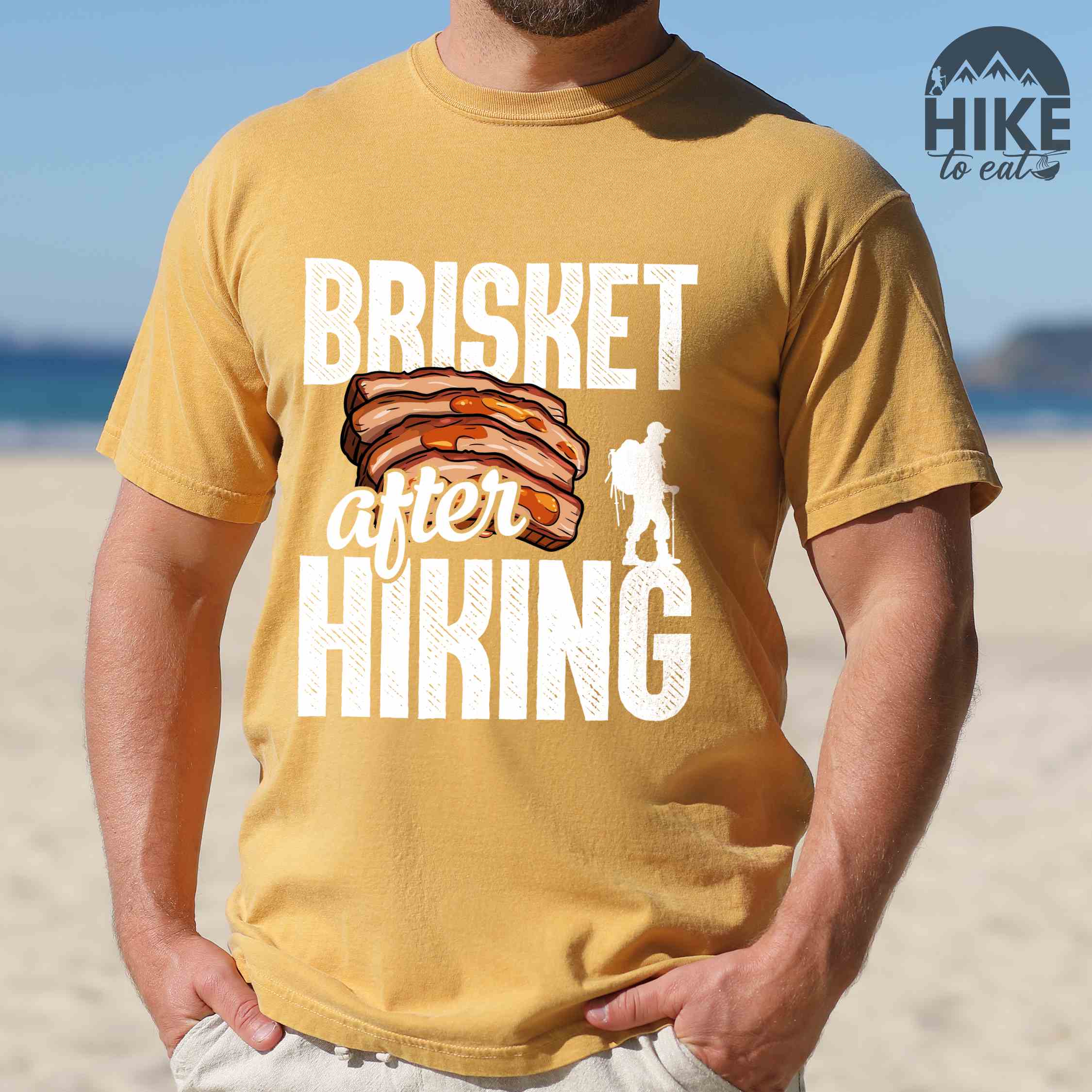 Man wearing mustard "Brisket after hiking" colored vintage tee with bold typography, birds, mountains, illustrated bbq beef brisket, and backpacker for bbq lovers.