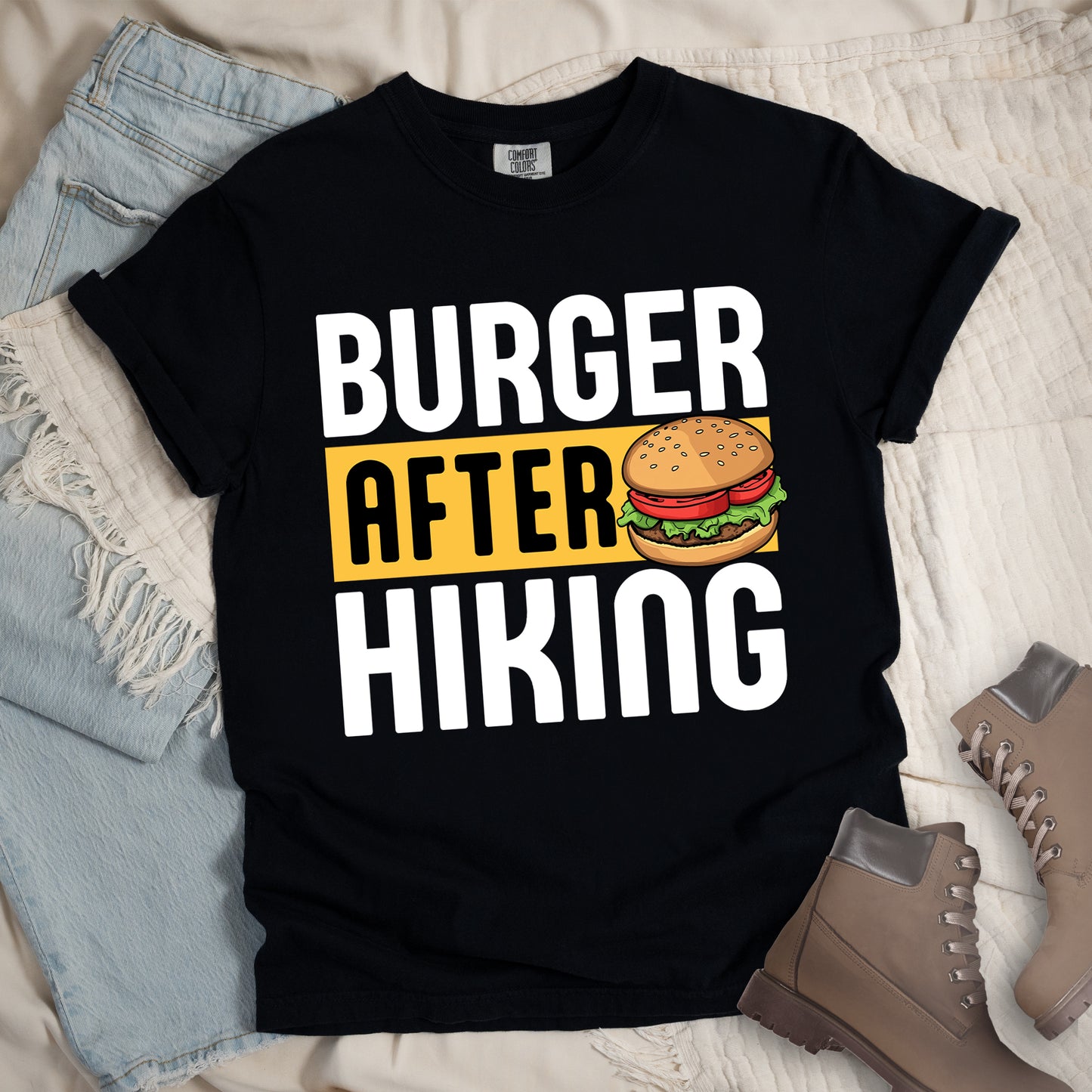 Black t-shirt with bold text reading "BURGER AFTER HIKING". "BURGER" appears in large white letters at the top, "AFTER" in black text on a yellow rectangular background in the middle, and "HIKING" in large white letters at the bottom. Features a detailed illustration of a hamburger with sesame seed bun, lettuce, tomato and patty next to the word "AFTER". The shirt features a classic crew neck and casual rolled sleeves, styled alongside light wash denim and brown hiking boots.