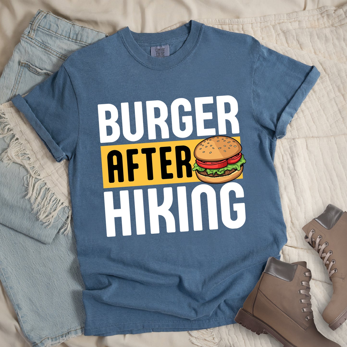 Blue Jean (Denim Blue) t-shirt with bold text reading "BURGER AFTER HIKING". "BURGER" appears in large white letters at the top, "AFTER" in black text on a yellow rectangular background in the middle, and "HIKING" in large white letters at the bottom. Features a detailed illustration of a hamburger with sesame seed bun, lettuce, tomato and patty next to the word "AFTER". The shirt features a classic crew neck and casual rolled sleeves, styled alongside light wash denim and brown hiking boots.