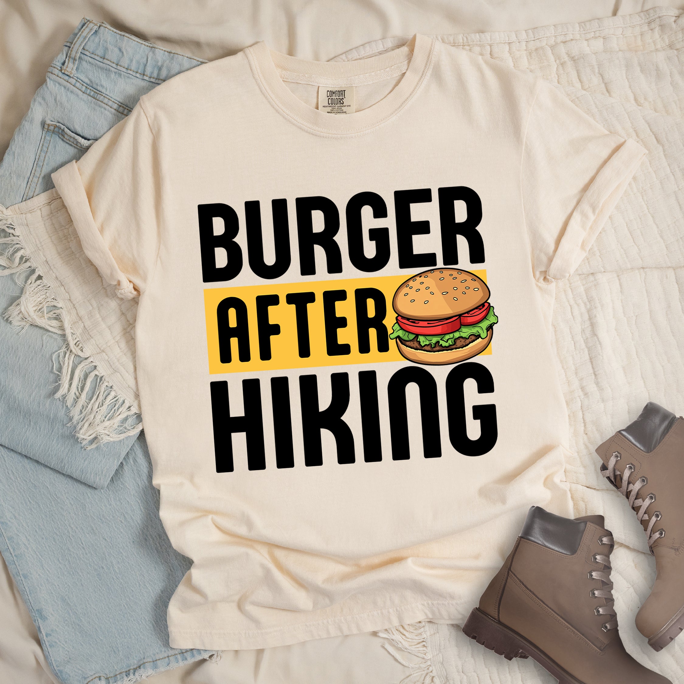 Ivory (Cream White) t-shirt with bold text reading "BURGER AFTER HIKING". "BURGER" appears in large black letters at the top, "AFTER" in black text on a yellow rectangular background in the middle, and "HIKING" in large black letters at the bottom. Features a detailed illustration of a hamburger with sesame seed bun, lettuce, tomato and patty next to the word "AFTER". The shirt features a classic crew neck and casual rolled sleeves, styled alongside light wash denim and brown hiking boots.