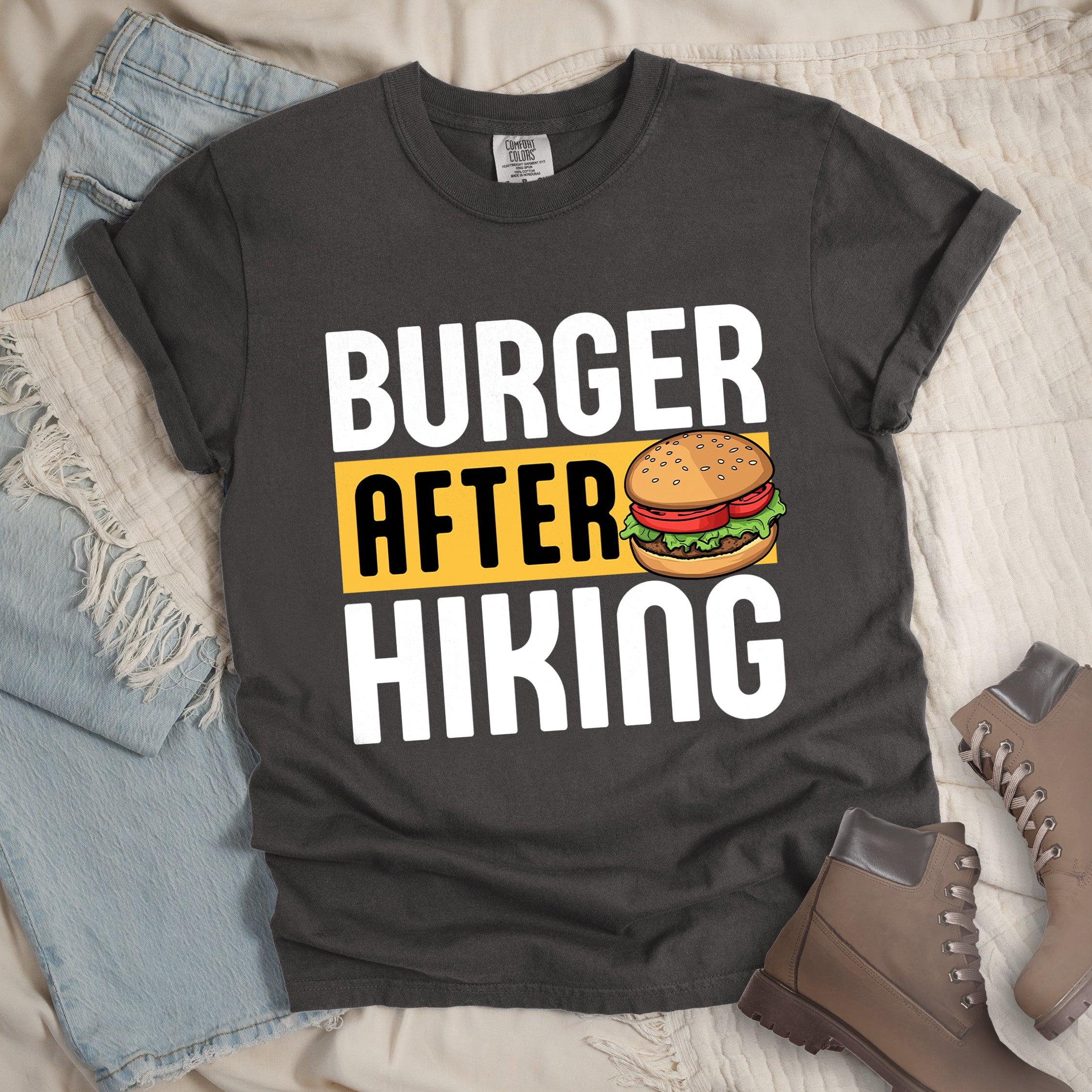 Pepper (Dark Gray) t-shirt with bold text reading "BURGER AFTER HIKING". "BURGER" appears in large white letters at the top, "AFTER" in black text on a yellow rectangular background in the middle, and "HIKING" in large white letters at the bottom. Features a detailed illustration of a hamburger with sesame seed bun, lettuce, tomato and patty next to the word "AFTER". The shirt features a classic crew neck and casual rolled sleeves, styled alongside light wash denim and brown hiking boots.