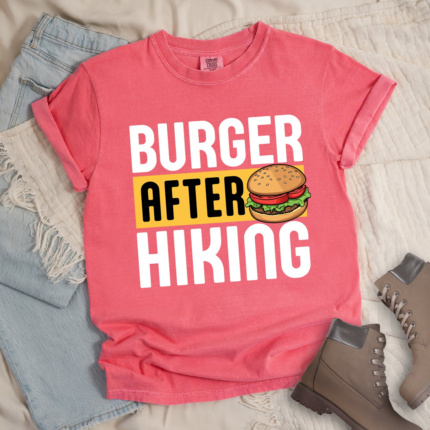 Watermelon (Light Coral) t-shirt with bold text reading "BURGER AFTER HIKING". "BURGER" appears in large white letters at the top, "AFTER" in black text on a yellow rectangular background in the middle, and "HIKING" in large white letters at the bottom. Features a detailed illustration of a hamburger with sesame seed bun, lettuce, tomato and patty next to the word "AFTER". The shirt features a classic crew neck and casual rolled sleeves, styled alongside light wash denim and brown hiking boots.