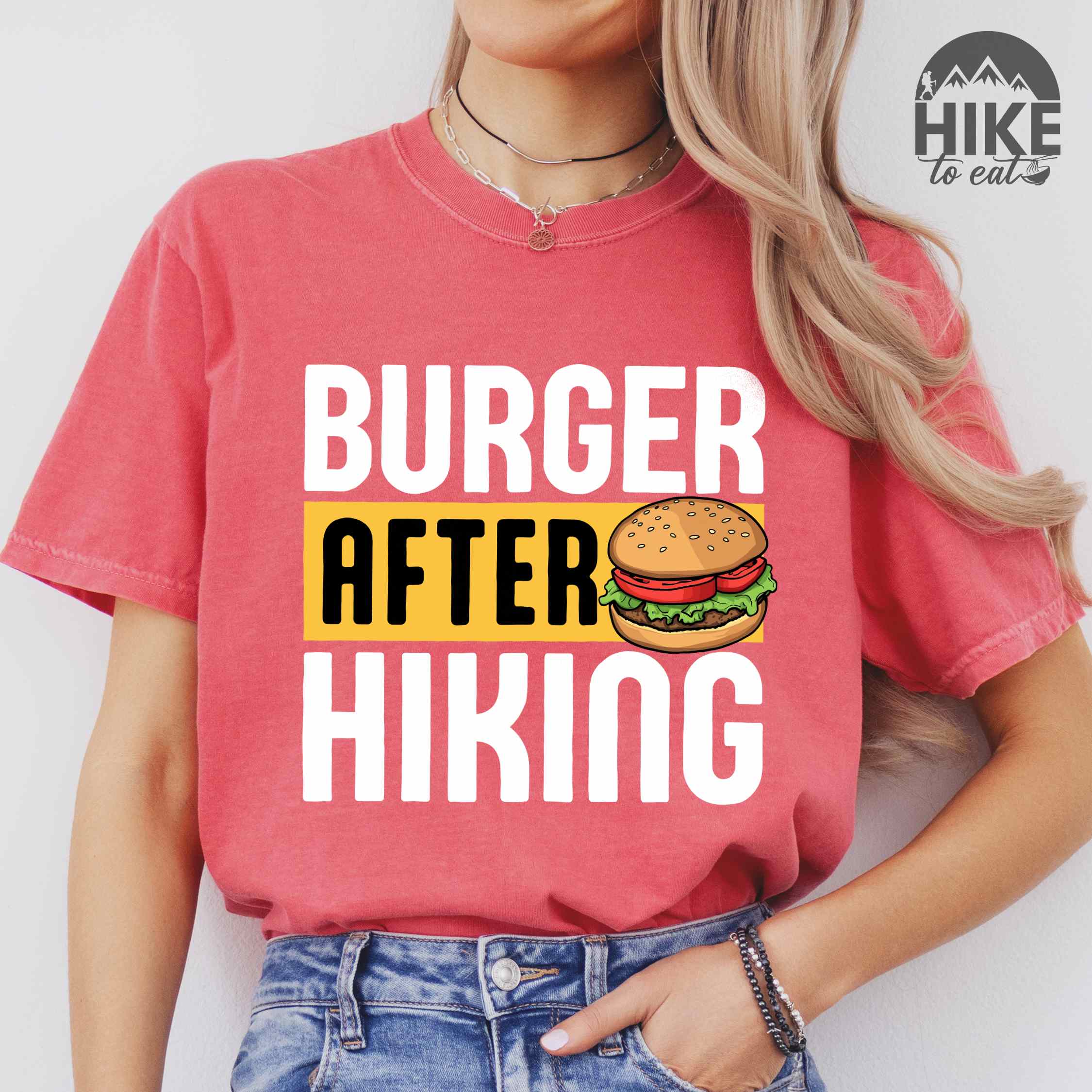 Woman wearing watermelon "Burger after Hiking" colored vintage tee with bold typography, birds, mountains, illustrated juicy hamburger, and backpacker for burger enthusiasts.