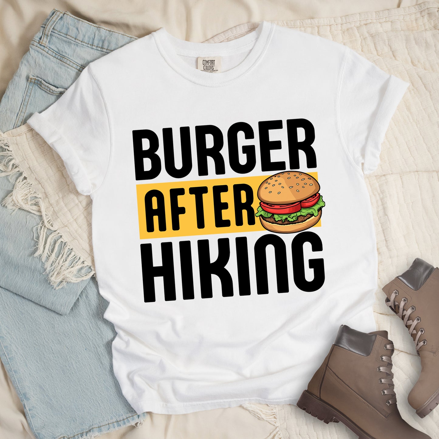 White t-shirt with bold text reading "BURGER AFTER HIKING". "BURGER" appears in large black letters at the top, "AFTER" in black text on a yellow rectangular background in the middle, and "HIKING" in large black letters at the bottom. Features a detailed illustration of a hamburger with sesame seed bun, lettuce, tomato and patty next to the word "AFTER". The shirt features a classic crew neck and casual rolled sleeves, styled alongside light wash denim and brown hiking boots.