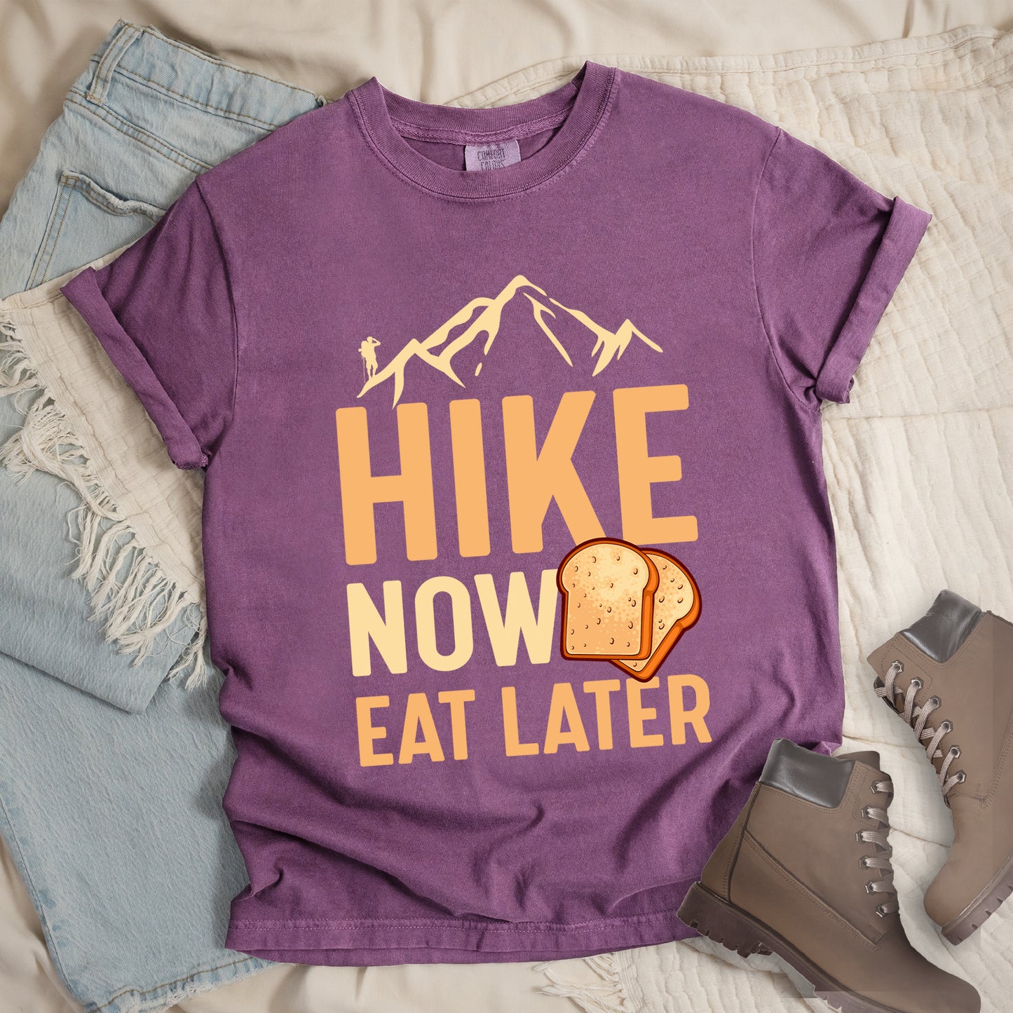 A Berry (Dark Violet) t-shirt with a design showing "HIKE NOW EAT LATER" in gradient orange and tan text. Above "HIKE" is a mountain silhouette with a small hiker figure. A sandwich/toast illustration appears between "NOW" and "EAT LATER". The text is arranged in three lines with "HIKE" at the top in largest font, followed by "NOW" and "EAT LATER" below. The shirt features a classic crew neck and casual rolled sleeves, styled alongside light wash denim and brown hiking boots.
