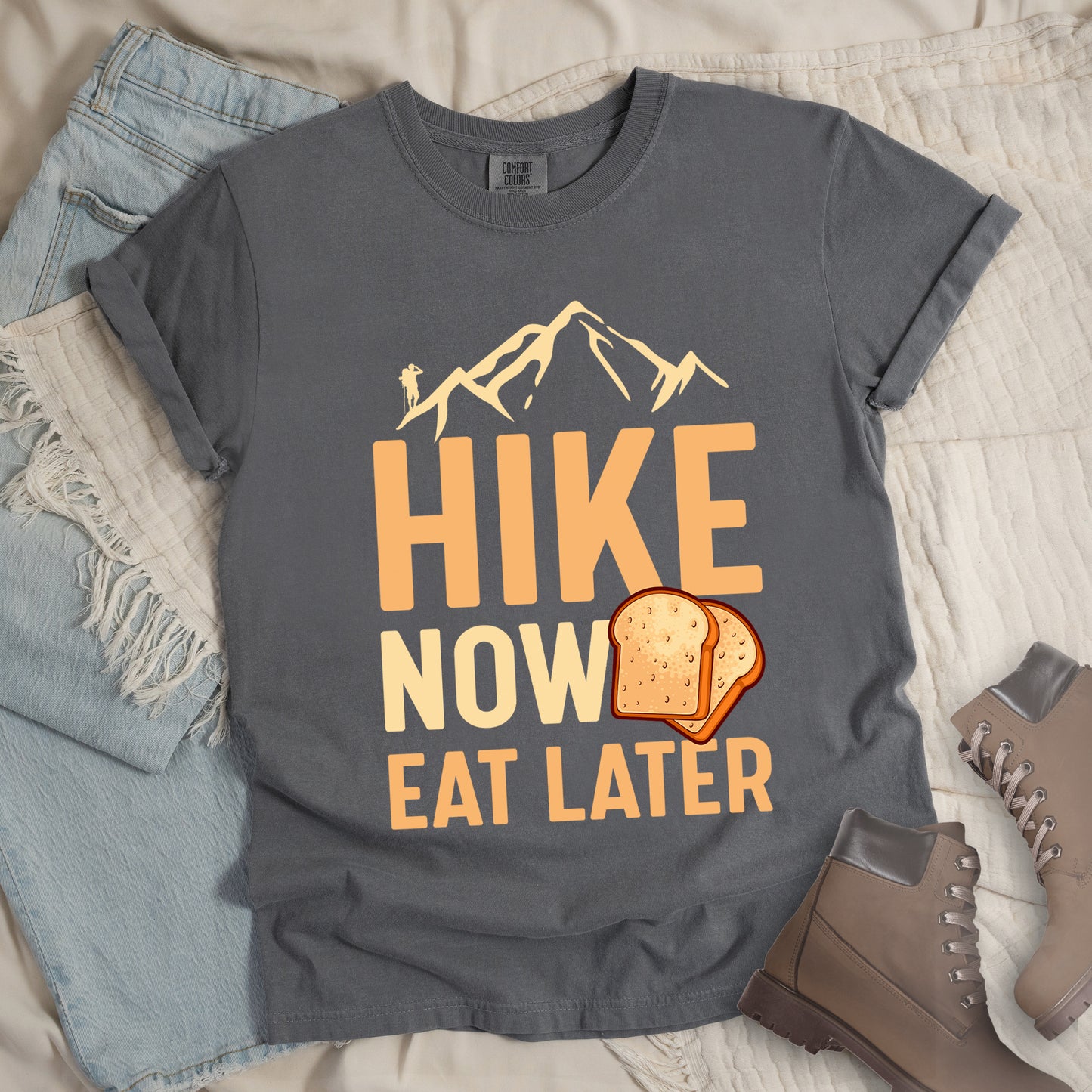 A grey t-shirt with a design showing "HIKE NOW EAT LATER" in gradient orange and tan text. Above "HIKE" is a mountain silhouette with a small hiker figure. A sandwich/toast illustration appears between "NOW" and "EAT LATER". The text is arranged in three lines with "HIKE" at the top in largest font, followed by "NOW" and "EAT LATER" below. The shirt features a classic crew neck and casual rolled sleeves, styled alongside light wash denim and brown hiking boots.