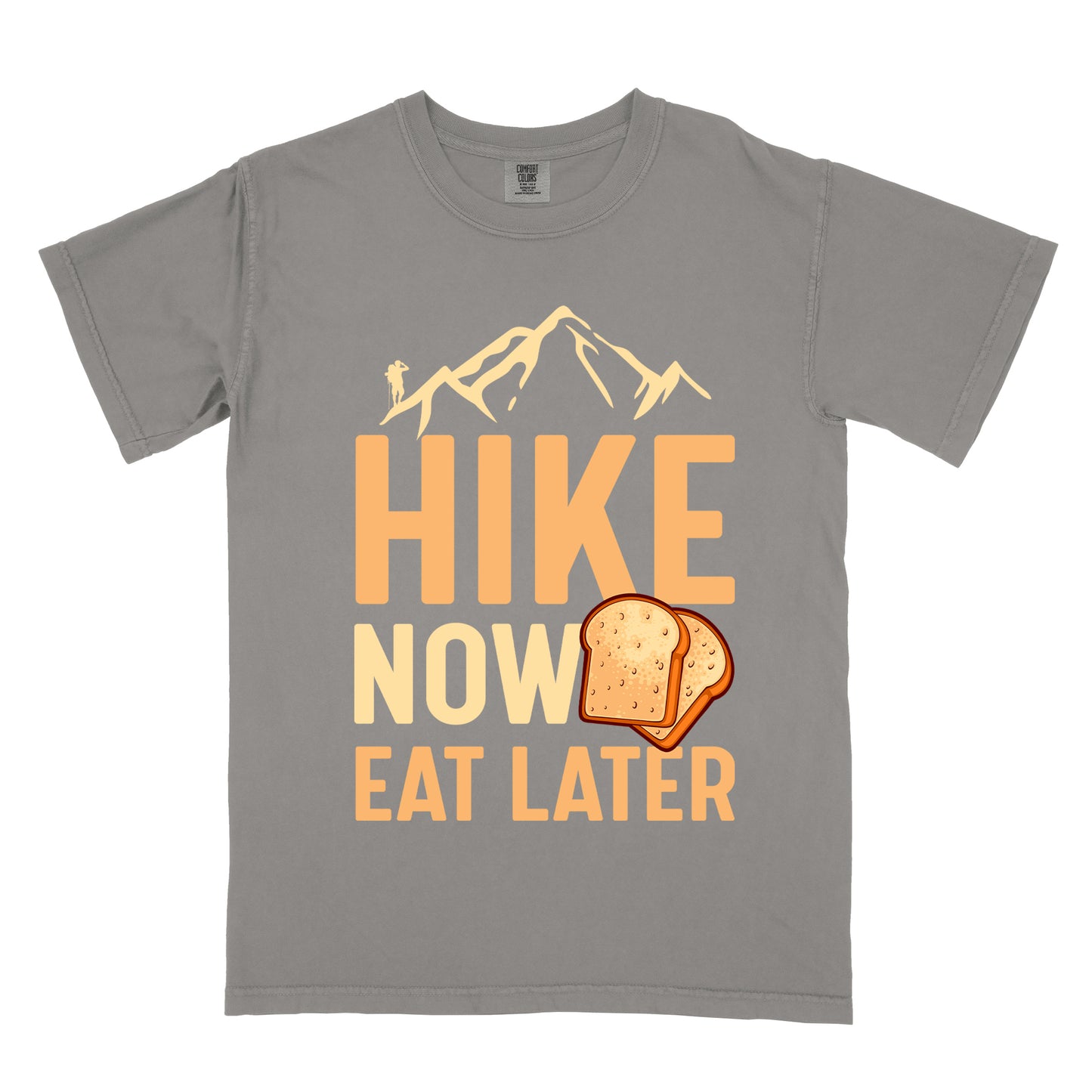 A grey t-shirt with a design showing "HIKE NOW EAT LATER" in gradient orange and tan text. Above "HIKE" is a mountain silhouette with a small hiker figure. A sandwich/toast illustration appears between "NOW" and "EAT LATER". The text is arranged in three lines with "HIKE" at the top in largest font, followed by "NOW" and "EAT LATER" below.