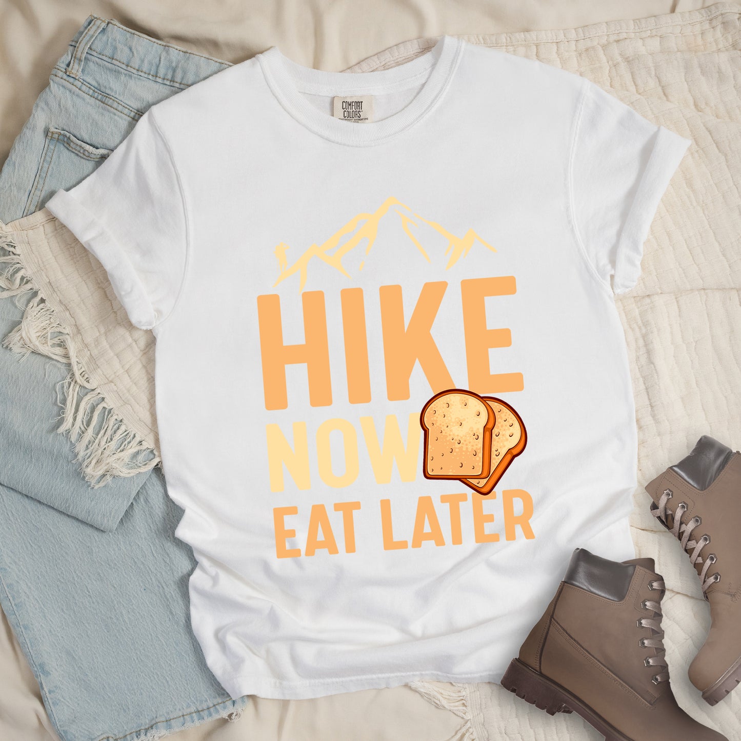 A white t-shirt with a design showing "HIKE NOW EAT LATER" in gradient orange and tan text. Above "HIKE" is a mountain silhouette with a small hiker figure. A sandwich/toast illustration appears between "NOW" and "EAT LATER". The text is arranged in three lines with "HIKE" at the top in largest font, followed by "NOW" and "EAT LATER" below. The shirt features a classic crew neck and casual rolled sleeves, styled alongside light wash denim and brown hiking boots.
