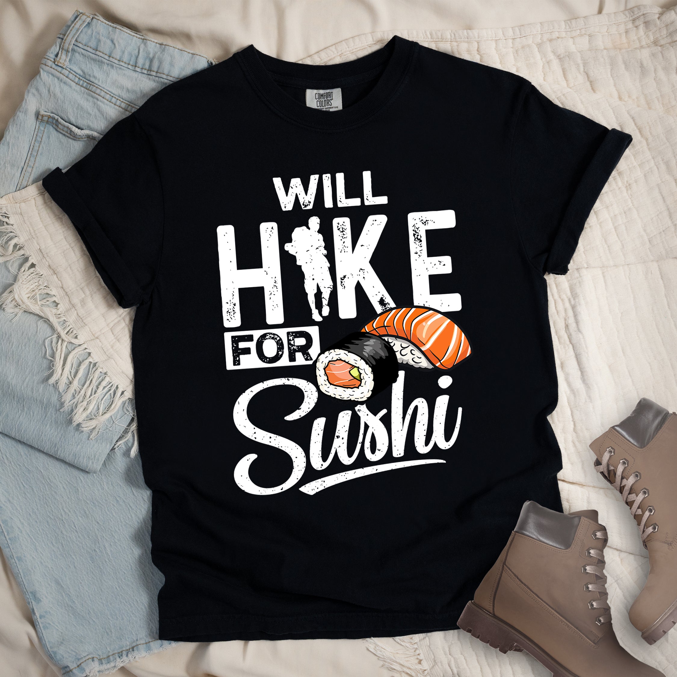 Black t-shirt with white distressed text reading "WILL HIKE FOR Sushi". "WILL" appears at the top, "HIKE" in large letters with a hiker silhouette integrated into the "I", "FOR" in smaller text, and "Sushi" in stylized script at the bottom. Features colorful sushi illustrations including a salmon nigiri and a maki roll. Features a classic crew neck and casual rolled sleeves, styled alongside light wash denim and brown hiking boots.