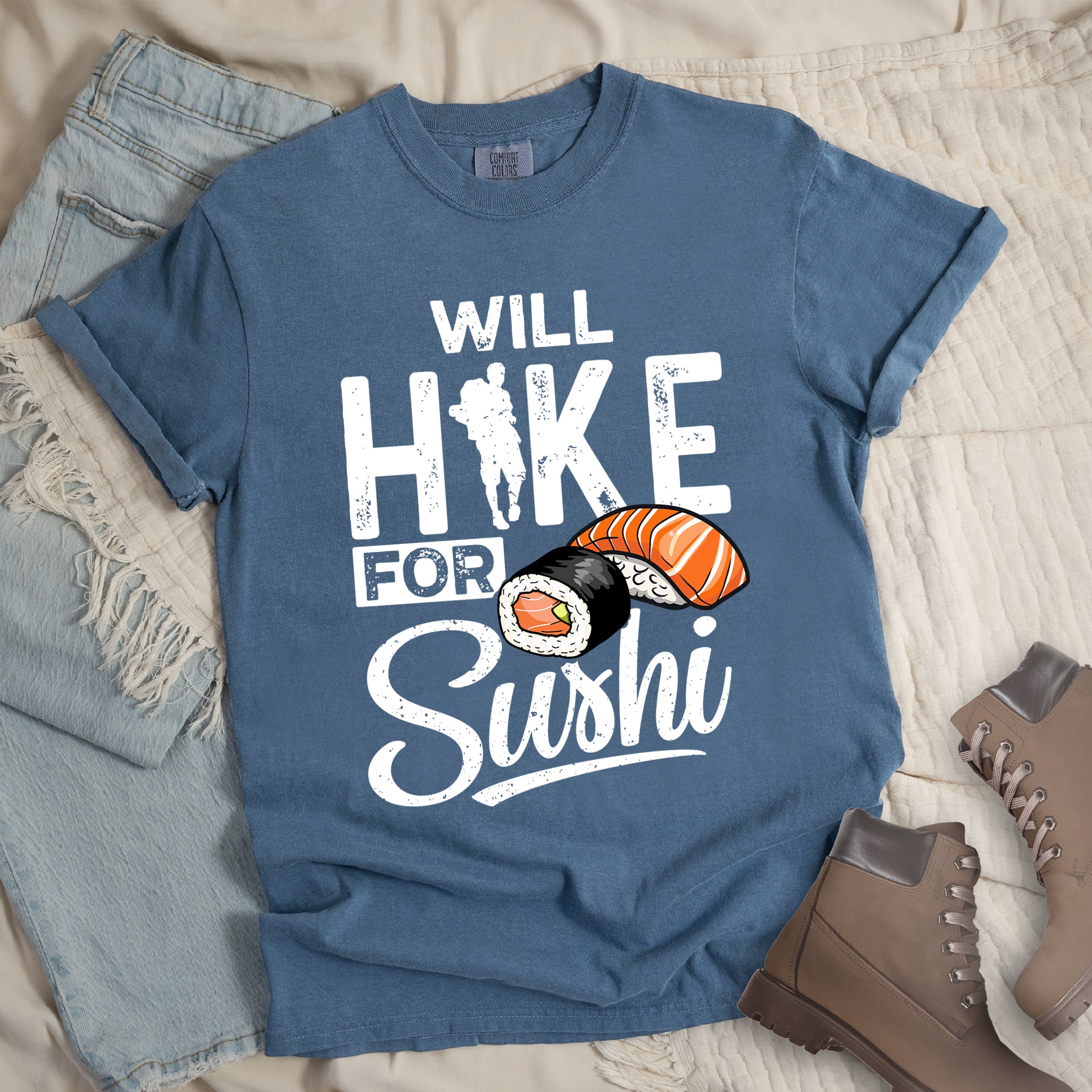 Blue Jean (Denim Blue) t-shirt with white distressed text reading "WILL HIKE FOR Sushi". "WILL" appears at the top, "HIKE" in large letters with a hiker silhouette integrated into the "I", "FOR" in smaller text, and "Sushi" in stylized script at the bottom. Features colorful sushi illustrations including a salmon nigiri and a maki roll. Features a classic crew neck and casual rolled sleeves, styled alongside light wash denim and brown hiking boots.