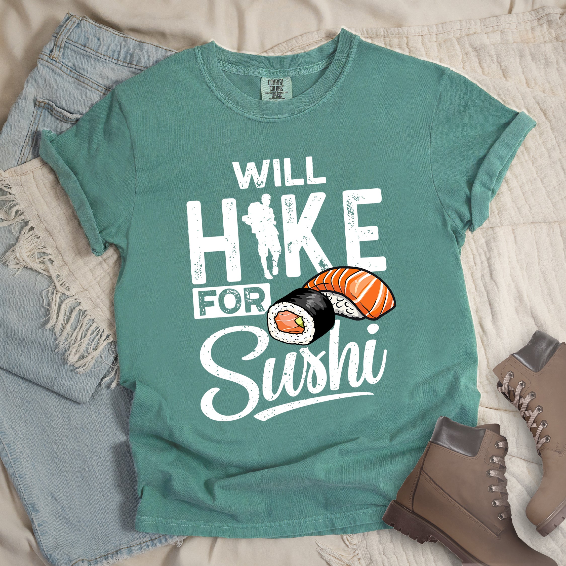 Light Green t-shirt with white distressed text reading "WILL HIKE FOR Sushi". "WILL" appears at the top, "HIKE" in large letters with a hiker silhouette integrated into the "I", "FOR" in smaller text, and "Sushi" in stylized script at the bottom. Features colorful sushi illustrations including a salmon nigiri and a maki roll. Features a classic crew neck and casual rolled sleeves, styled alongside light wash denim and brown hiking boots.