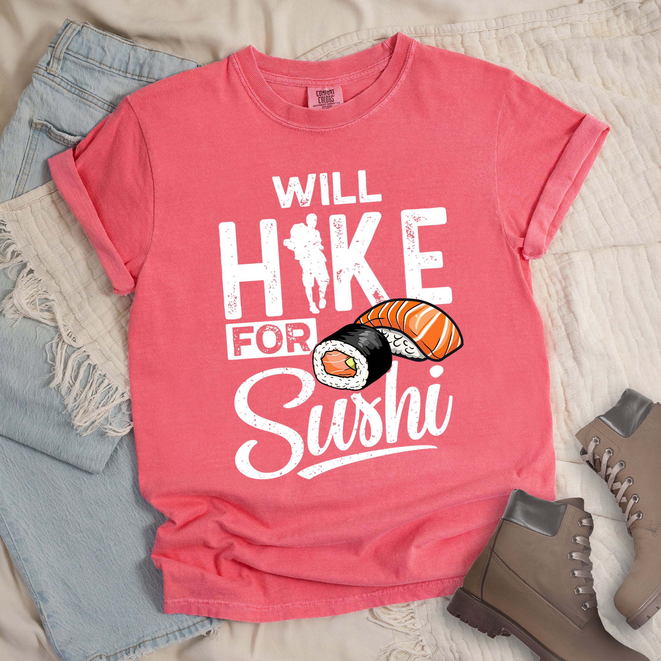 Watermelon (Light Coral) t-shirt with white distressed text reading "WILL HIKE FOR Sushi". "WILL" appears at the top, "HIKE" in large letters with a hiker silhouette integrated into the "I", "FOR" in smaller text, and "Sushi" in stylized script at the bottom. Features colorful sushi illustrations including a salmon nigiri and a maki roll. Features a classic crew neck and casual rolled sleeves, styled alongside light wash denim and brown hiking boots.