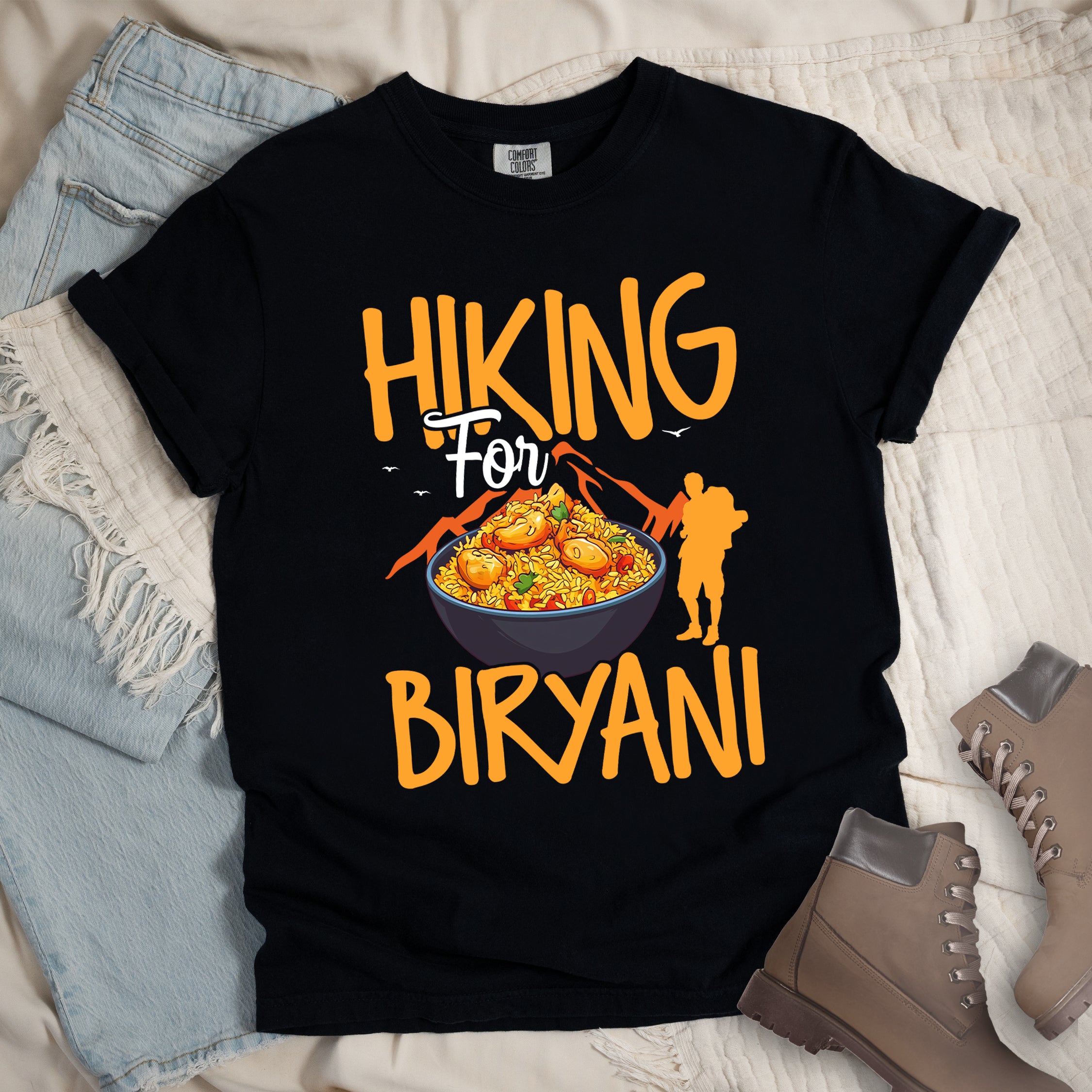 Black t-shirt with orange text reading "HIKING For BIRYANI". "HIKING" appears in large orange letters at the top, "For" in white script in the middle, and "BIRYANI" in orange at the bottom. Features a detailed illustration of a blue bowl filled with yellow biryani rice and meat pieces in the center, bright orange mountain silhouettes in the background, and an orange hiker silhouette on the right side.