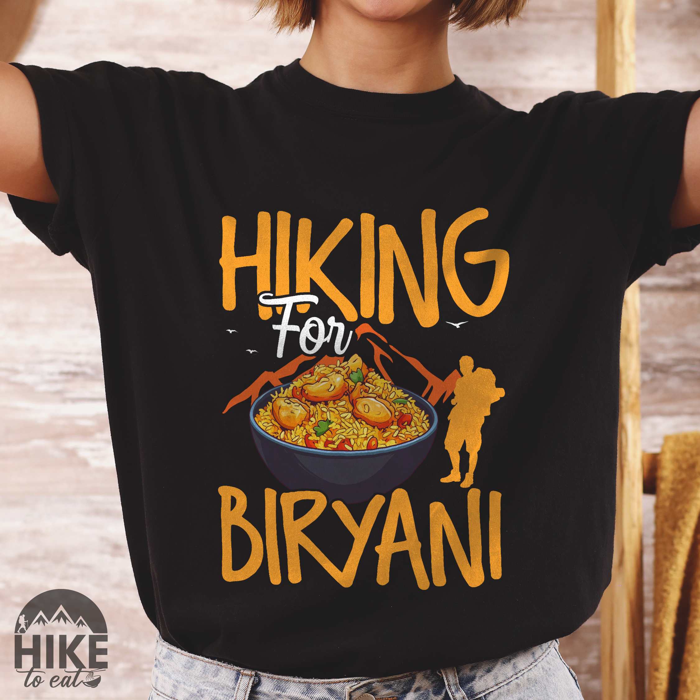 Woman wearing black "Hiking for Biryani" colored vintage tee with bold typography, birds, mountains, illustrated bowl of biryani rice, and backpacker for rice lovers.