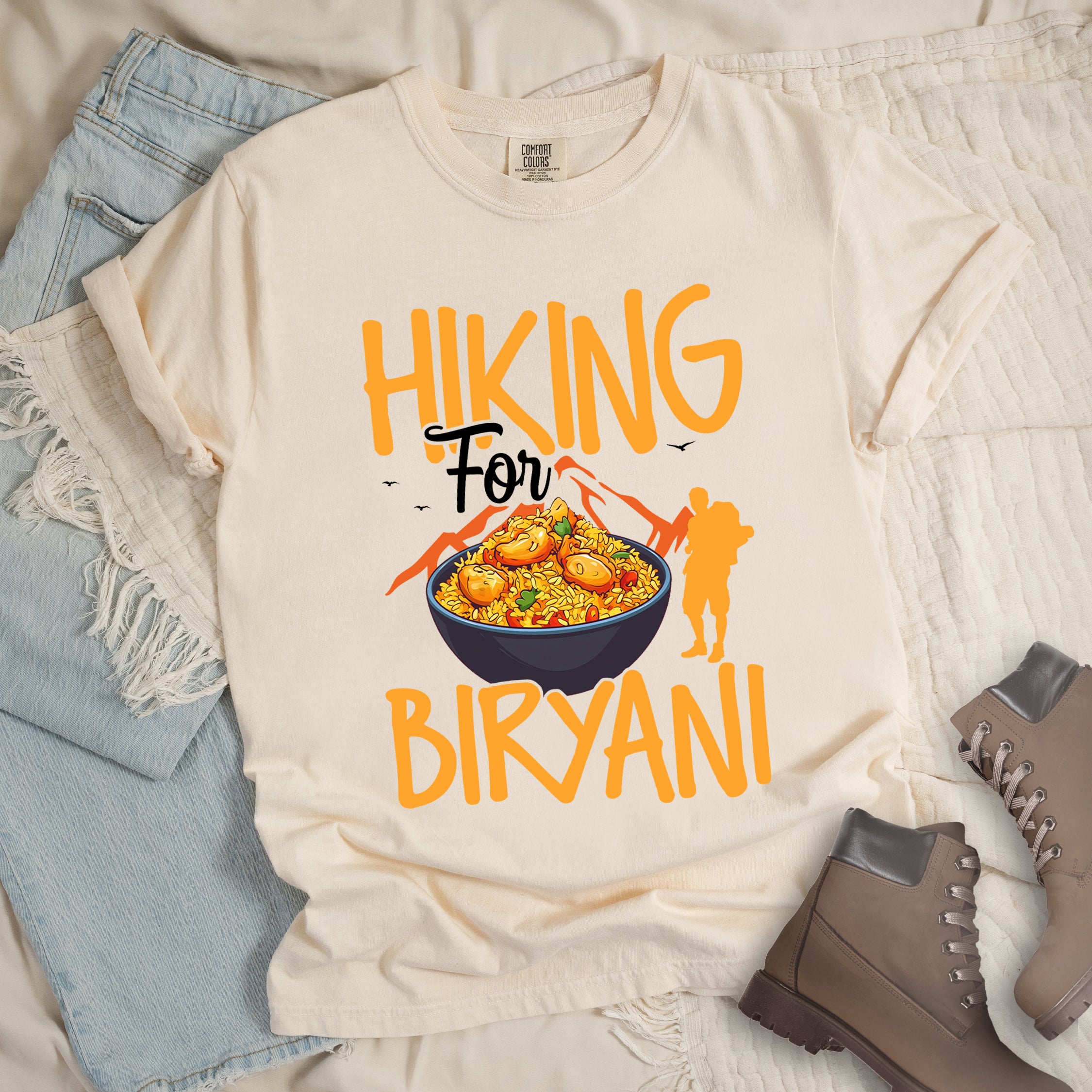 Ivory (Cream White) t-shirt with orange text reading "HIKING For BIRYANI". "HIKING" appears in large orange letters at the top, "For" in black script in the middle, and "BIRYANI" in orange at the bottom. Features a detailed illustration of a blue bowl filled with yellow biryani rice and meat pieces in the center, bright orange mountain silhouettes in the background, and an orange hiker silhouette on the right side.