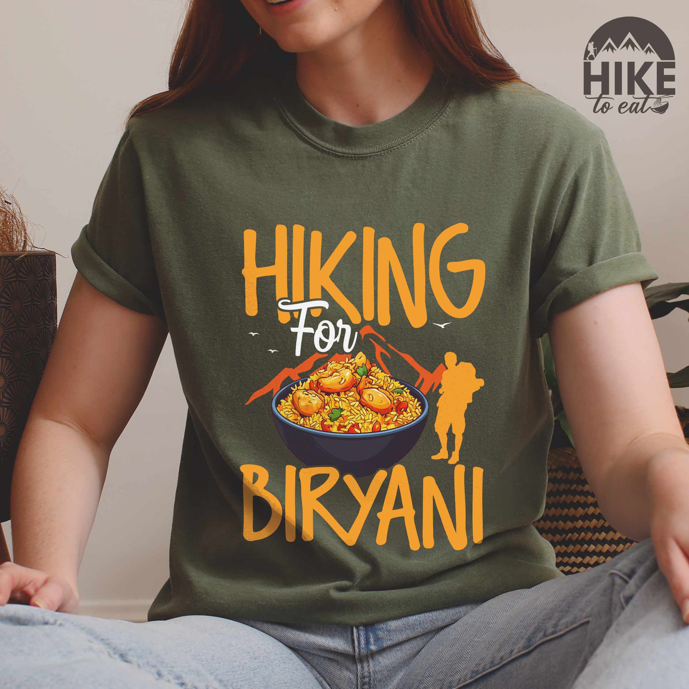 Woman wearing moss "Hiking for Biryani" colored vintage tee with bold typography, birds, mountains, illustrated bowl of biryani rice, and backpacker for rice lovers.