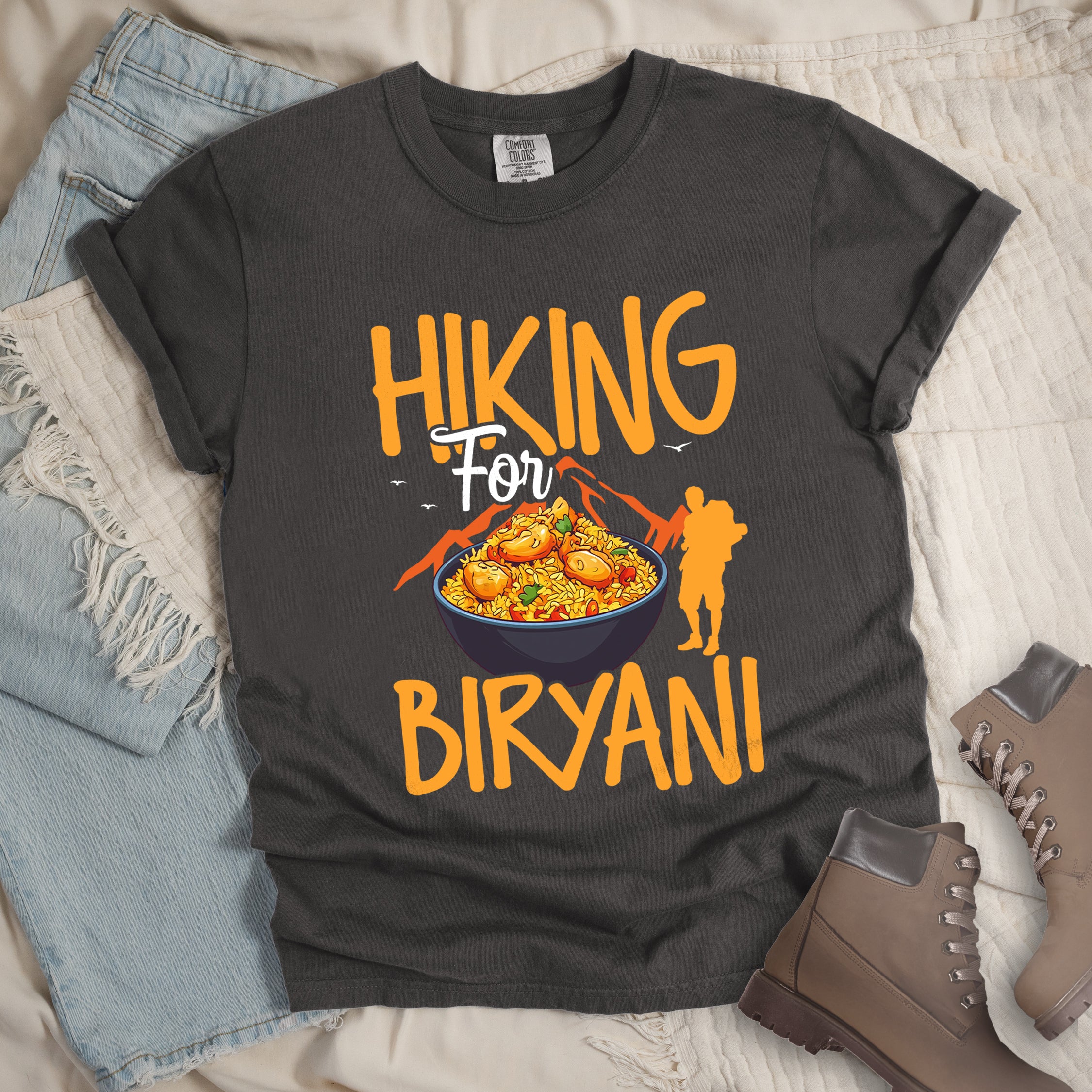 Pepper (Dark Gray) t-shirt with orange text reading "HIKING For BIRYANI". "HIKING" appears in large orange letters at the top, "For" in white script in the middle, and "BIRYANI" in orange at the bottom. Features a detailed illustration of a blue bowl filled with yellow biryani rice and meat pieces in the center, bright orange mountain silhouettes in the background, and an orange hiker silhouette on the right side.