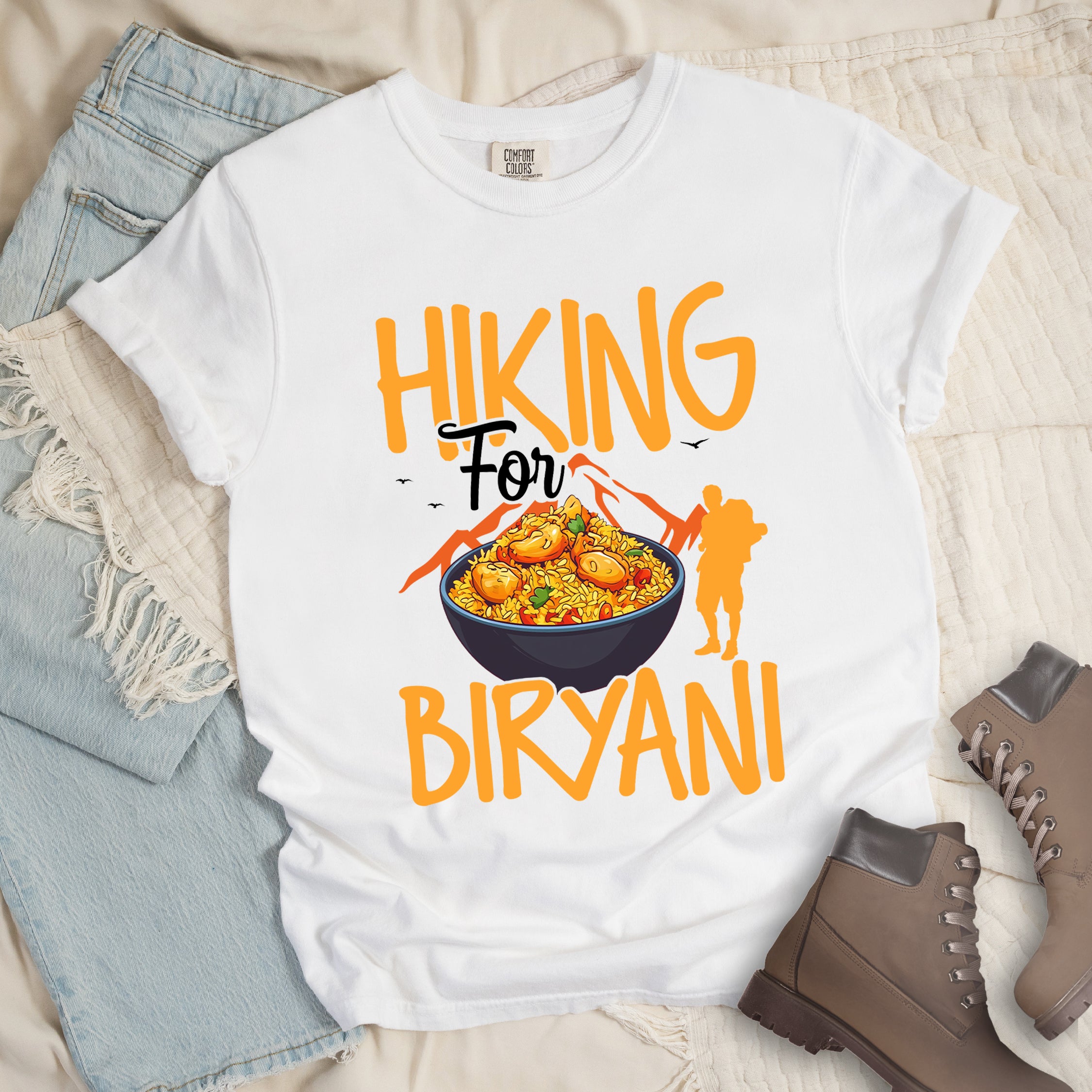 White t-shirt with orange text reading "HIKING For BIRYANI". "HIKING" appears in large orange letters at the top, "For" in black script in the middle, and "BIRYANI" in orange at the bottom. Features a detailed illustration of a blue bowl filled with yellow biryani rice and meat pieces in the center, bright orange mountain silhouettes in the background, and an orange hiker silhouette on the right side.