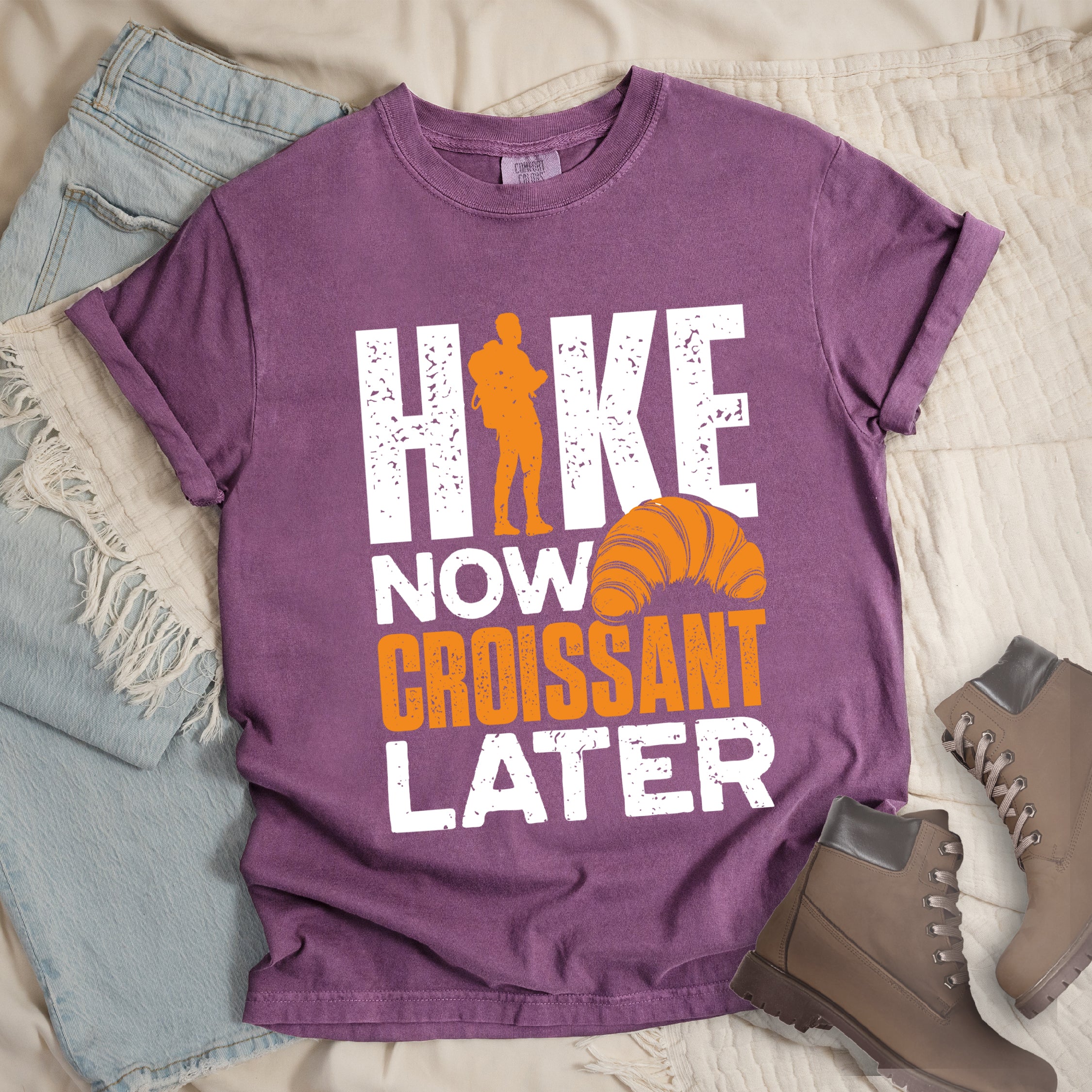 Berry (Dark Violet) t-shirt with white and orange distressed text reading "HIKE NOW CROISSANT LATER". "HIKE" appears in large white letters at the top with an orange hiker silhouette replacing the letter "I", "NOW" in white in the middle, "CROISSANT" in orange, and "LATER" in white at the bottom. Features an illustration of an orange croissant between "NOW" and "LATER". Features a classic crew neck and casual rolled sleeves, styled alongside light wash denim and brown hiking boots.