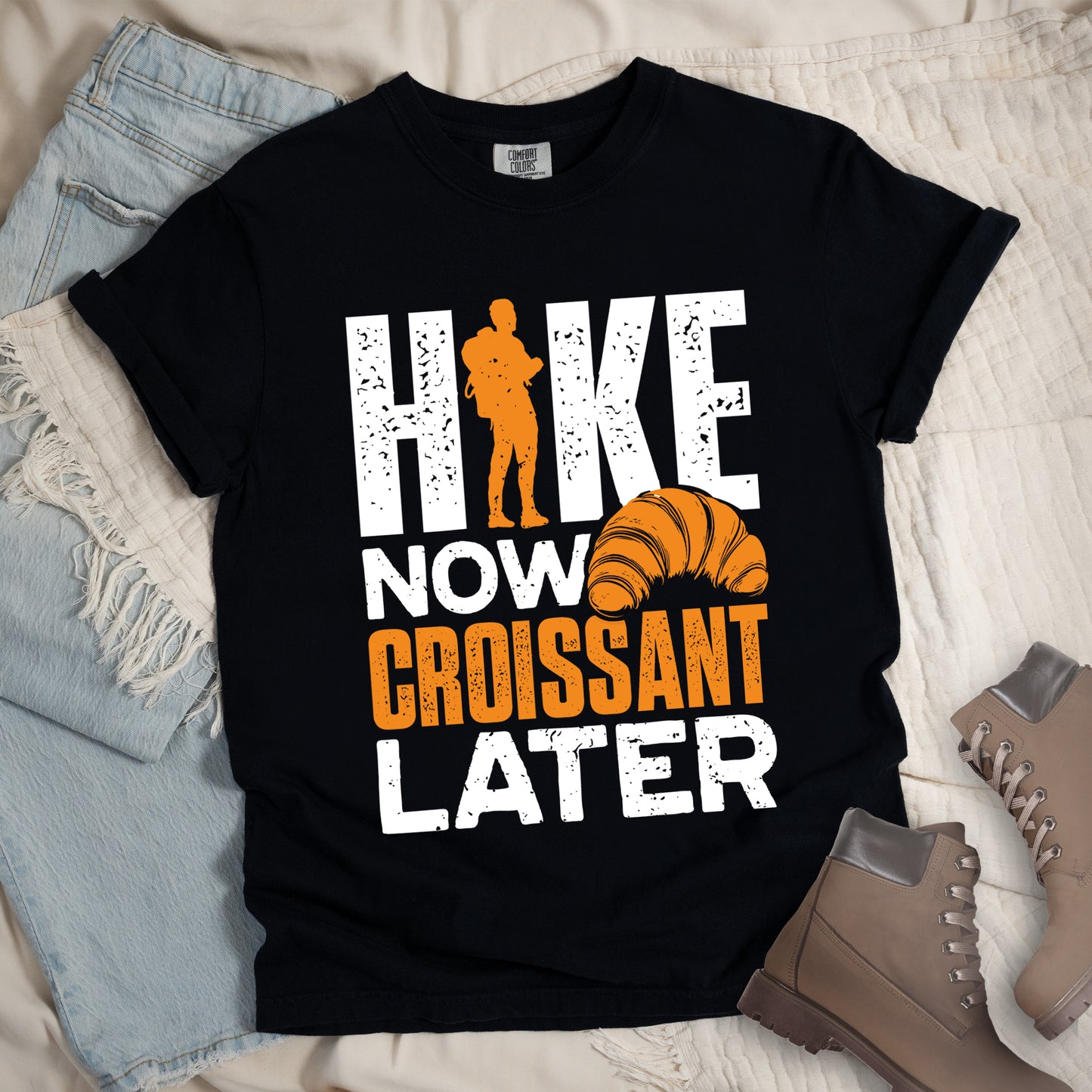 Black t-shirt with white and orange distressed text reading "HIKE NOW CROISSANT LATER". "HIKE" appears in large white letters at the top with an orange hiker silhouette replacing the letter "I", "NOW" in white in the middle, "CROISSANT" in orange, and "LATER" in white at the bottom. Features an illustration of an orange croissant between "NOW" and "LATER". Features a classic crew neck and casual rolled sleeves, styled alongside light wash denim and brown hiking boots.