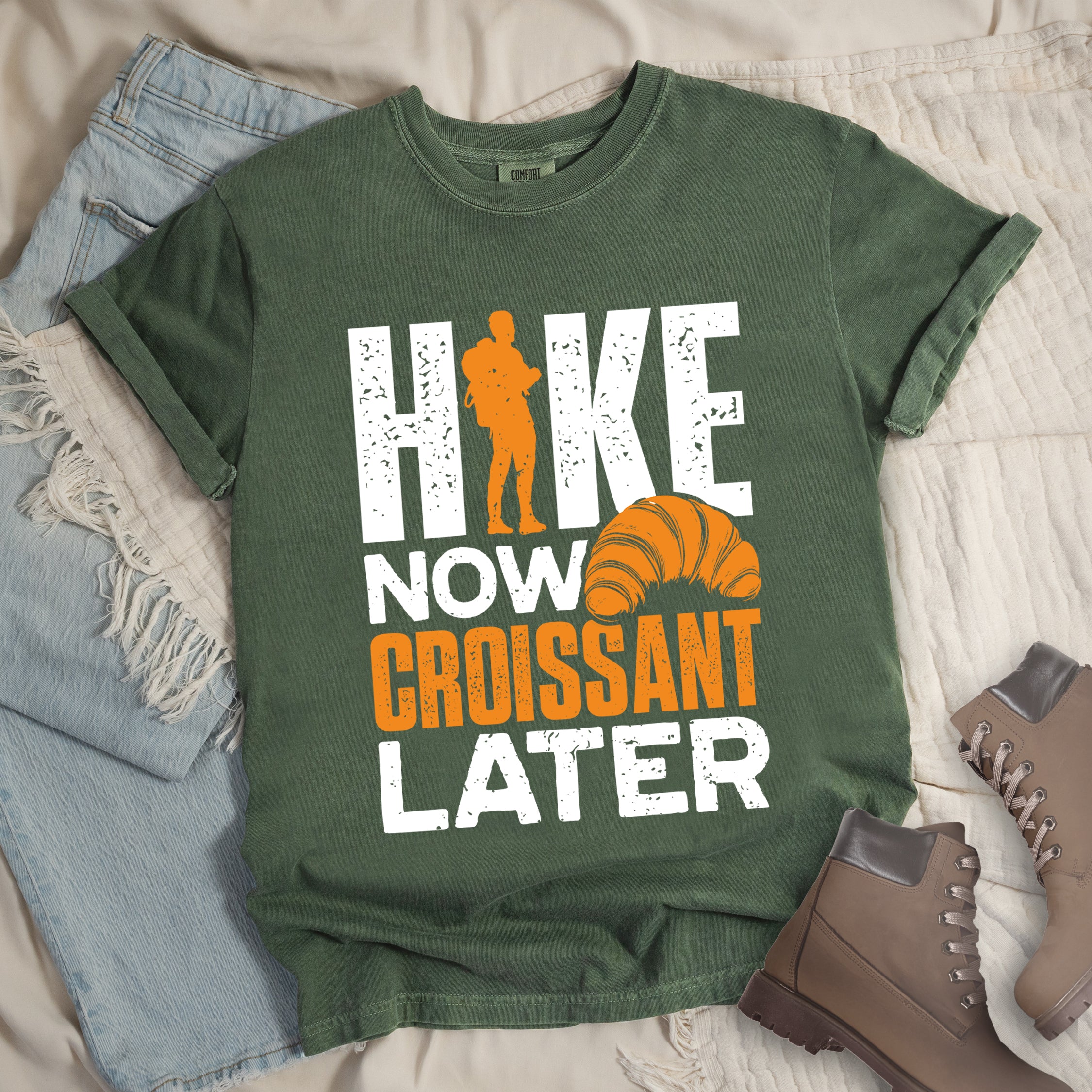 Moss (Dark Green) t-shirt with white and orange distressed text reading "HIKE NOW CROISSANT LATER". "HIKE" appears in large white letters at the top with an orange hiker silhouette replacing the letter "I", "NOW" in white in the middle, "CROISSANT" in orange, and "LATER" in white at the bottom. Features an illustration of an orange croissant between "NOW" and "LATER". Features a classic crew neck and casual rolled sleeves, styled alongside light wash denim and brown hiking boots.