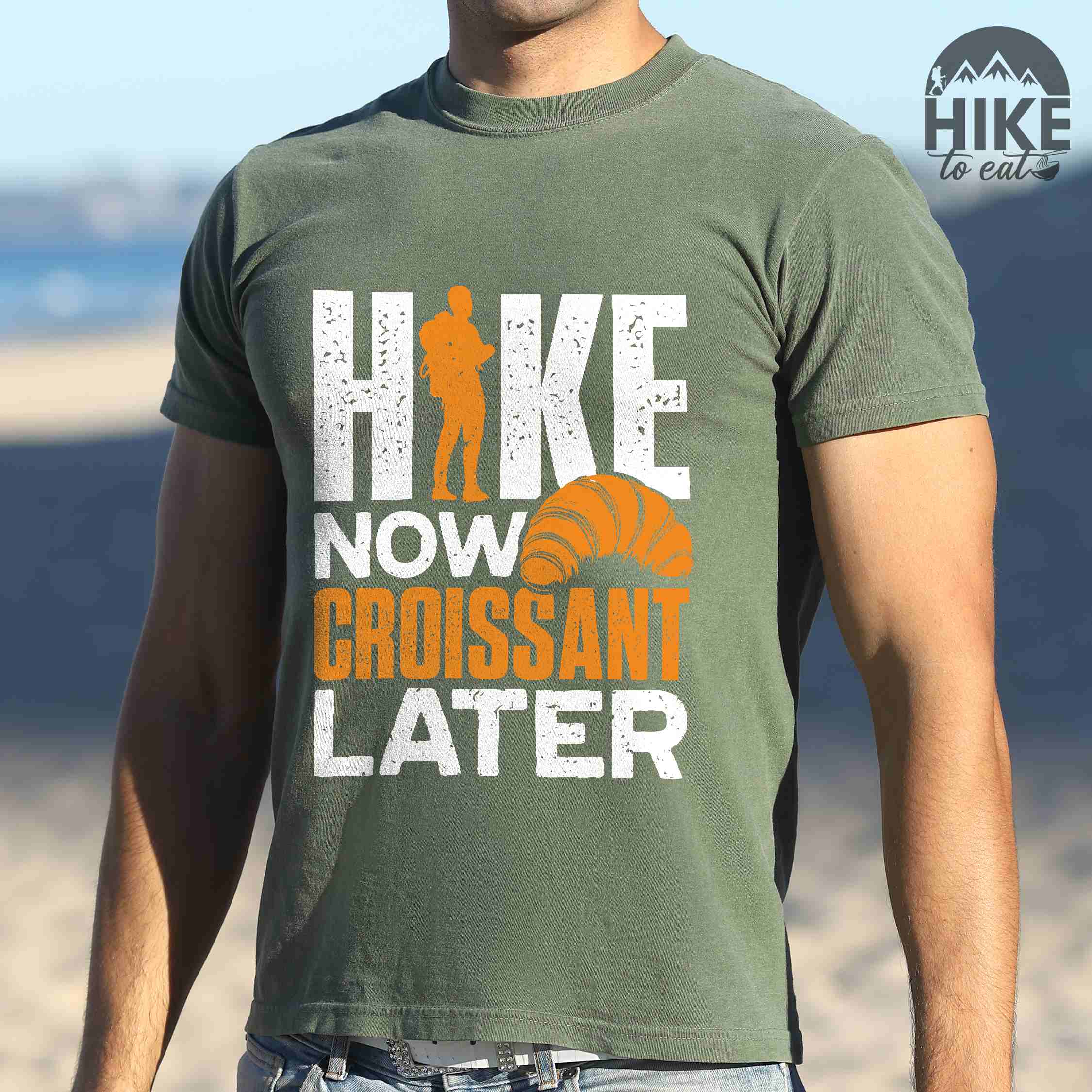 Man wearing moss "Hike now croissant later" colored vintage tee with bold typography, birds, mountains, illustrated baked croissant, and backpacker for pastry enthusiasts.