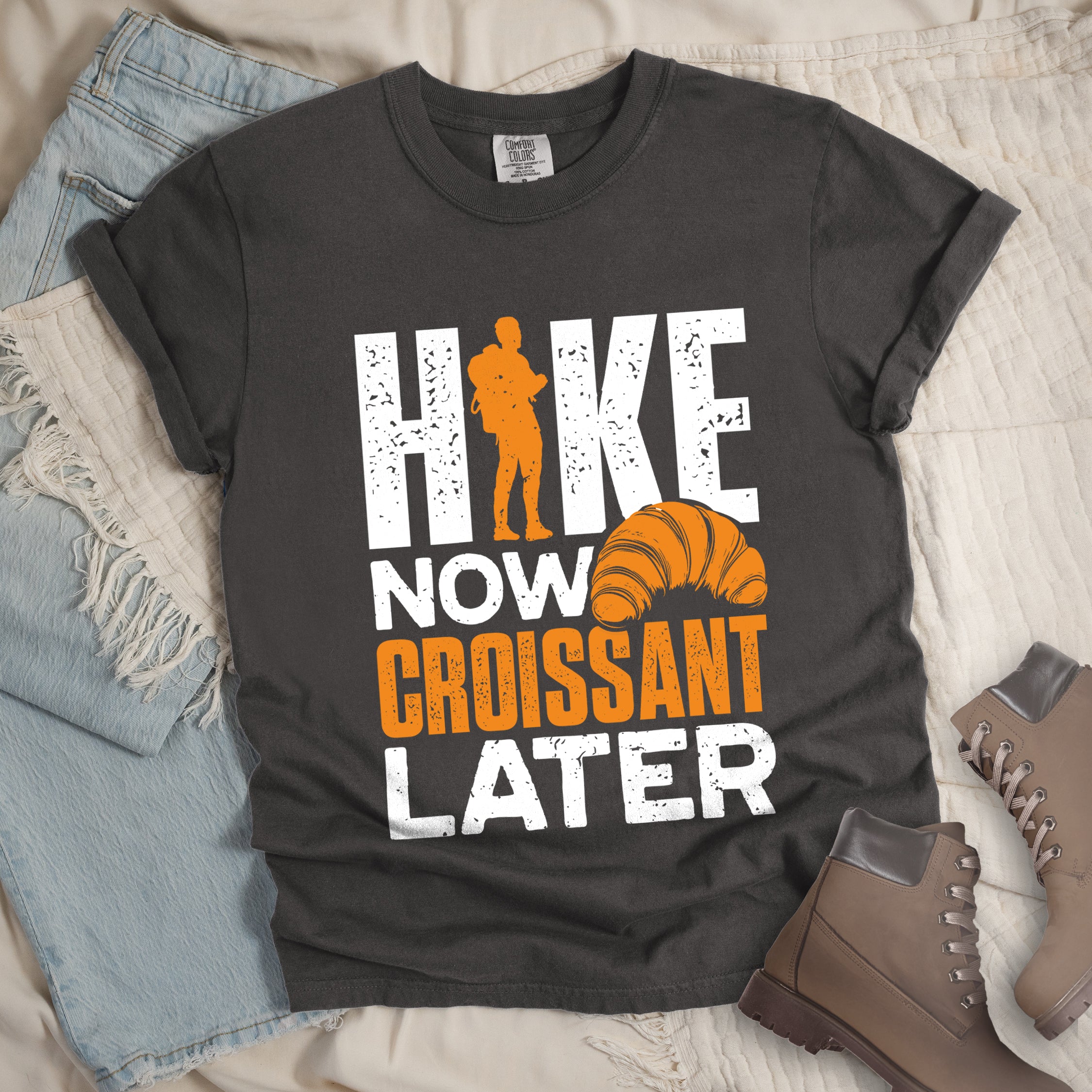 Pepper (Dark Gray) t-shirt with white and orange distressed text reading "HIKE NOW CROISSANT LATER". "HIKE" appears in large white letters at the top with an orange hiker silhouette replacing the letter "I", "NOW" in white in the middle, "CROISSANT" in orange, and "LATER" in white at the bottom. Features an illustration of an orange croissant between "NOW" and "LATER". Features a classic crew neck and casual rolled sleeves, styled alongside light wash denim and brown hiking boots.