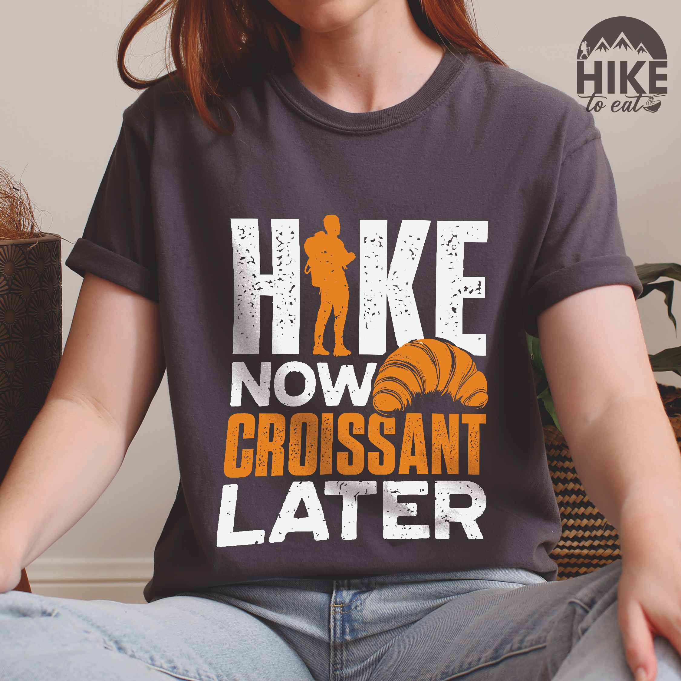 Woman wearing pepper "Hike now croissant later" colored vintage tee with bold typography, birds, mountains, illustrated baked croissant, and backpacker for pastry enthusiasts.