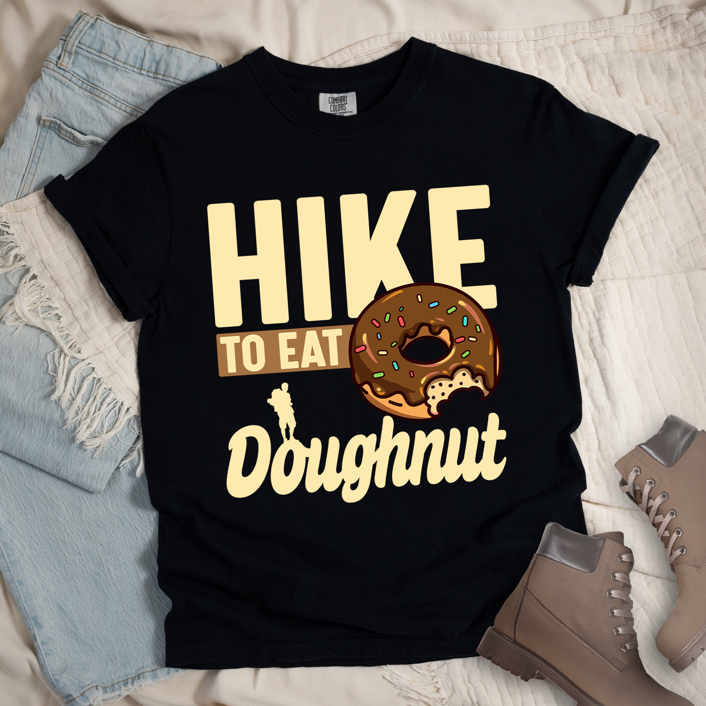 Black t-shirt with cream and tan text reading "HIKE TO EAT Doughnut". "HIKE" appears in large cream letters at the top, "TO EAT" on a tan rectangular background in the middle, and "Doughnut" in cream script at the bottom. Features a chocolate frosted doughnut with colorful sprinkles and a bite taken out of it on the right side, and a small cream silhouette of a hiker on the left.
