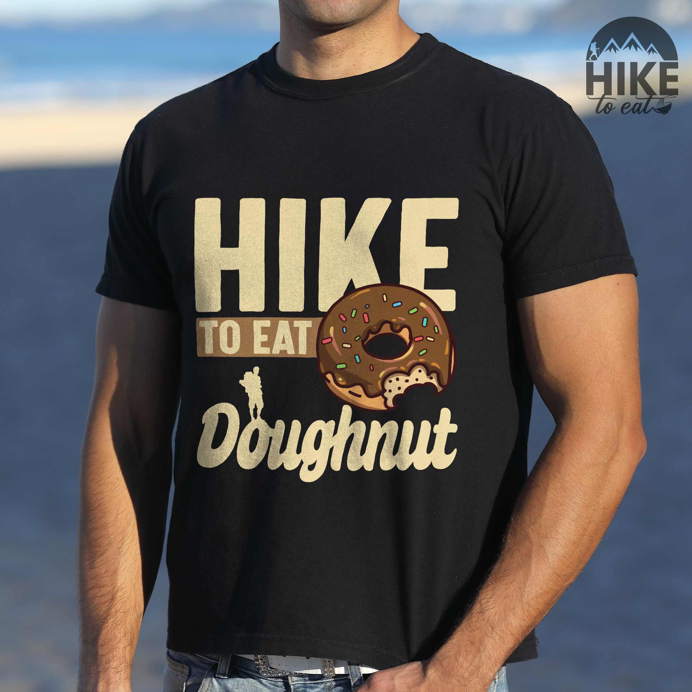 Man wearing black "Hike to eat doughnut" colored vintage tee with bold typography, birds, mountains, illustrated baked chocolate glazed donut, and backpacker for donut lovers.