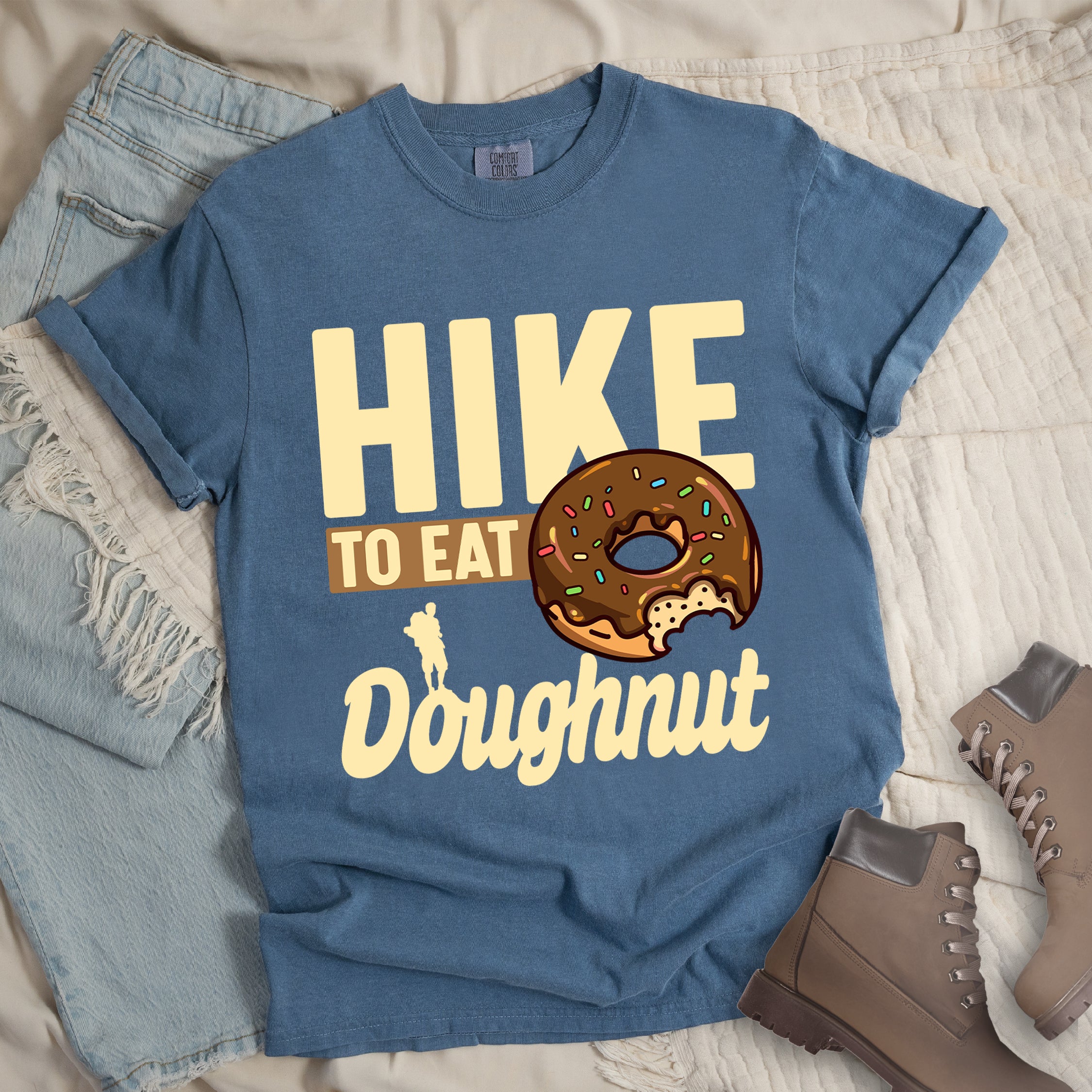 Blue Jean (Denim Blue) t-shirt with cream and tan text reading "HIKE TO EAT Doughnut". "HIKE" appears in large cream letters at the top, "TO EAT" on a tan rectangular background in the middle, and "Doughnut" in cream script at the bottom. Features a chocolate frosted doughnut with colorful sprinkles and a bite taken out of it on the right side, and a small cream silhouette of a hiker on the left. Features a classic crew neck and casual rolled sleeves, styled alongside light wash denim and brown hiking boots