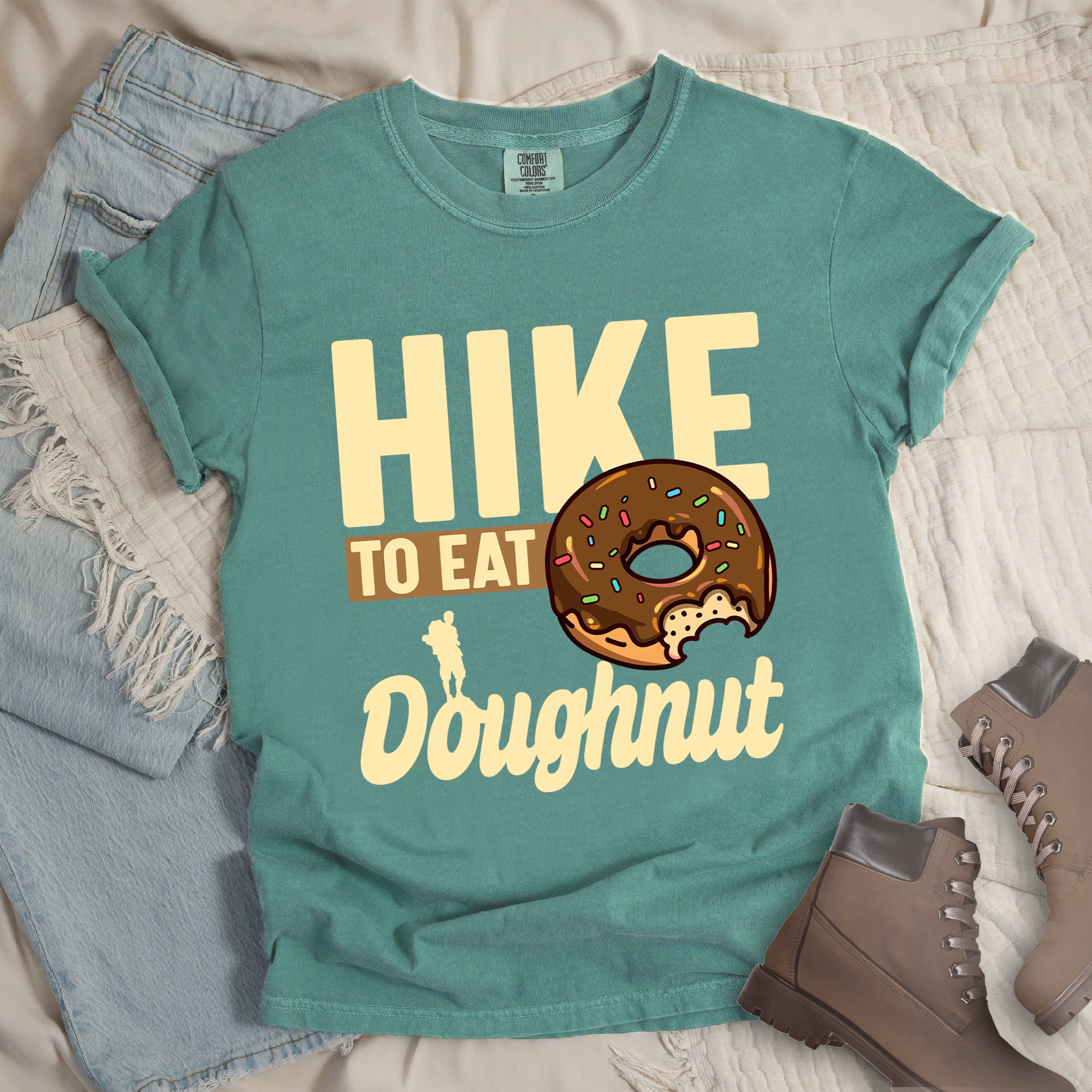 Light Green t-shirt with cream and tan text reading "HIKE TO EAT Doughnut". "HIKE" appears in large cream letters at the top, "TO EAT" on a tan rectangular background in the middle, and "Doughnut" in cream script at the bottom. Features a chocolate frosted doughnut with colorful sprinkles and a bite taken out of it on the right side, and a small cream silhouette of a hiker on the left.