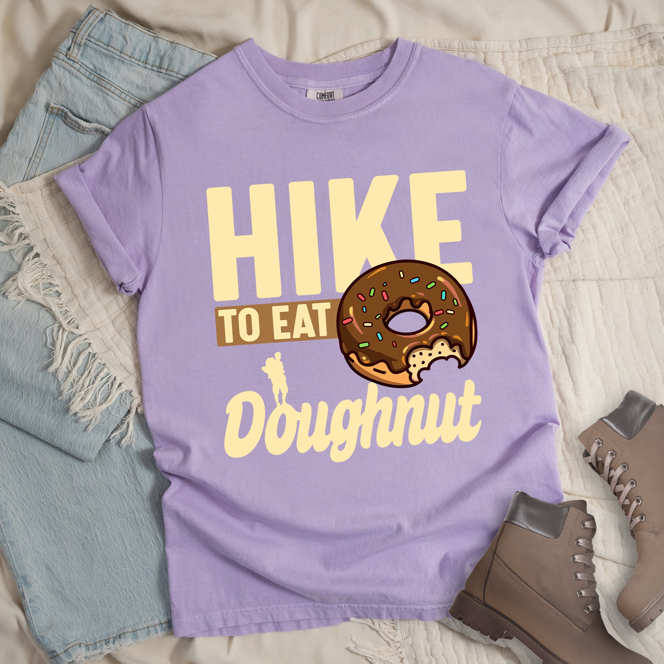 Orchid (Light Violet) t-shirt with cream and tan text reading "HIKE TO EAT Doughnut". "HIKE" appears in large cream letters at the top, "TO EAT" on a tan rectangular background in the middle, and "Doughnut" in cream script at the bottom. Features a chocolate frosted doughnut with colorful sprinkles and a bite taken out of it on the right side, and a small cream silhouette of a hiker on the left.