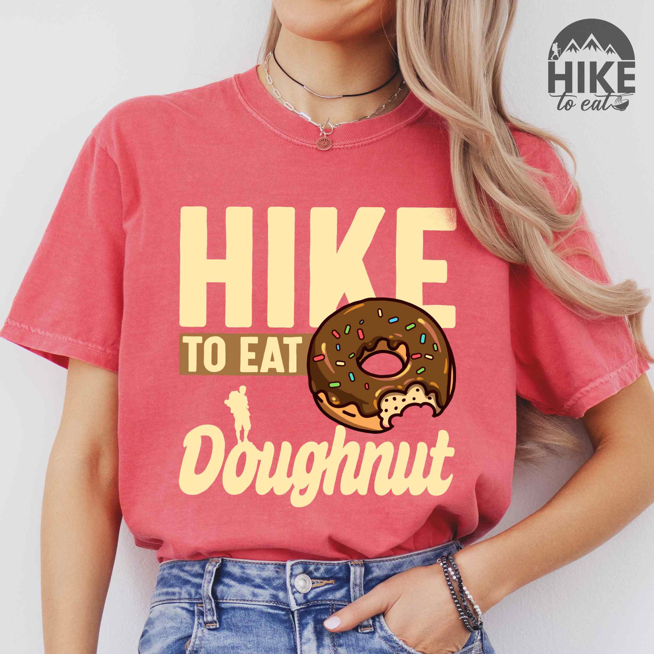 Woman wearing watermelon "Hike to eat doughnut" colored vintage tee with bold typography, birds, mountains, illustrated baked chocolate glazed donut, and backpacker for donut lovers.