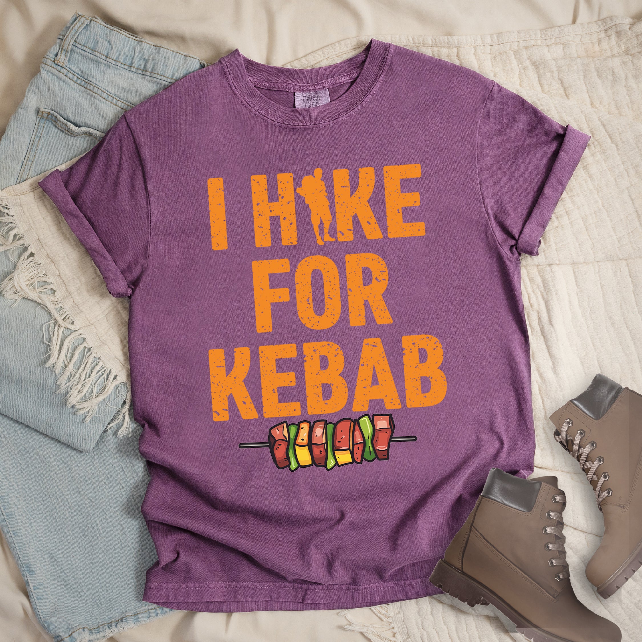 Berry (Dark Violet) t-shirt with orange distressed text reading "I HIKE FOR KEBAB". "I HIKE" appears at the top with a hiker silhouette integrated into the letter "I", "FOR" in the middle, and "KEBAB" at the bottom. Below the text is a colorful illustration of a kebab skewer with meat and vegetables. Features a classic crew neck and casual rolled sleeves, styled alongside light wash denim and brown hiking boots.