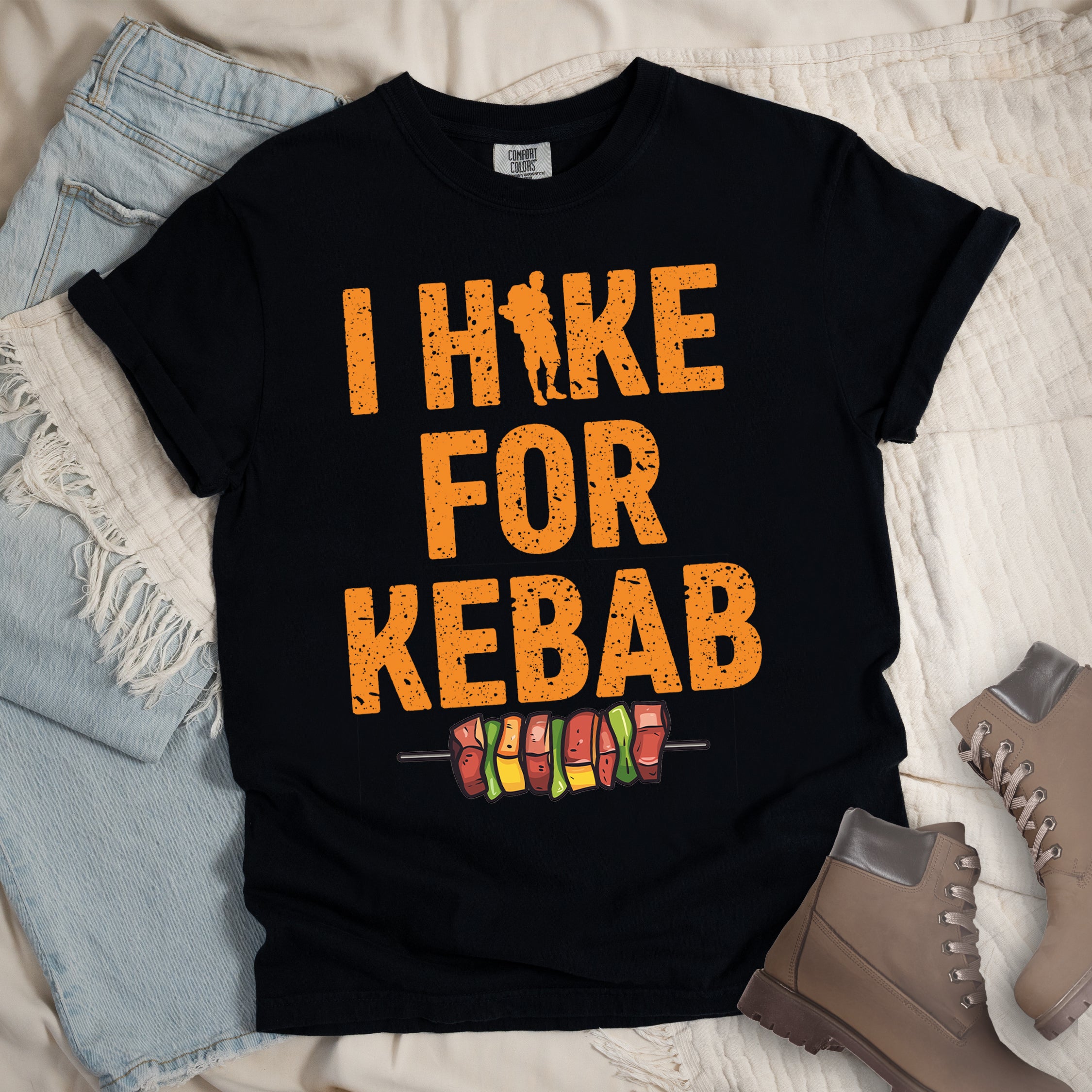 Black t-shirt with orange distressed text reading "I HIKE FOR KEBAB". "I HIKE" appears at the top with a hiker silhouette integrated into the letter "I", "FOR" in the middle, and "KEBAB" at the bottom. Below the text is a colorful illustration of a kebab skewer with meat and vegetables. Features a classic crew neck and casual rolled sleeves, styled alongside light wash denim and brown hiking boots.