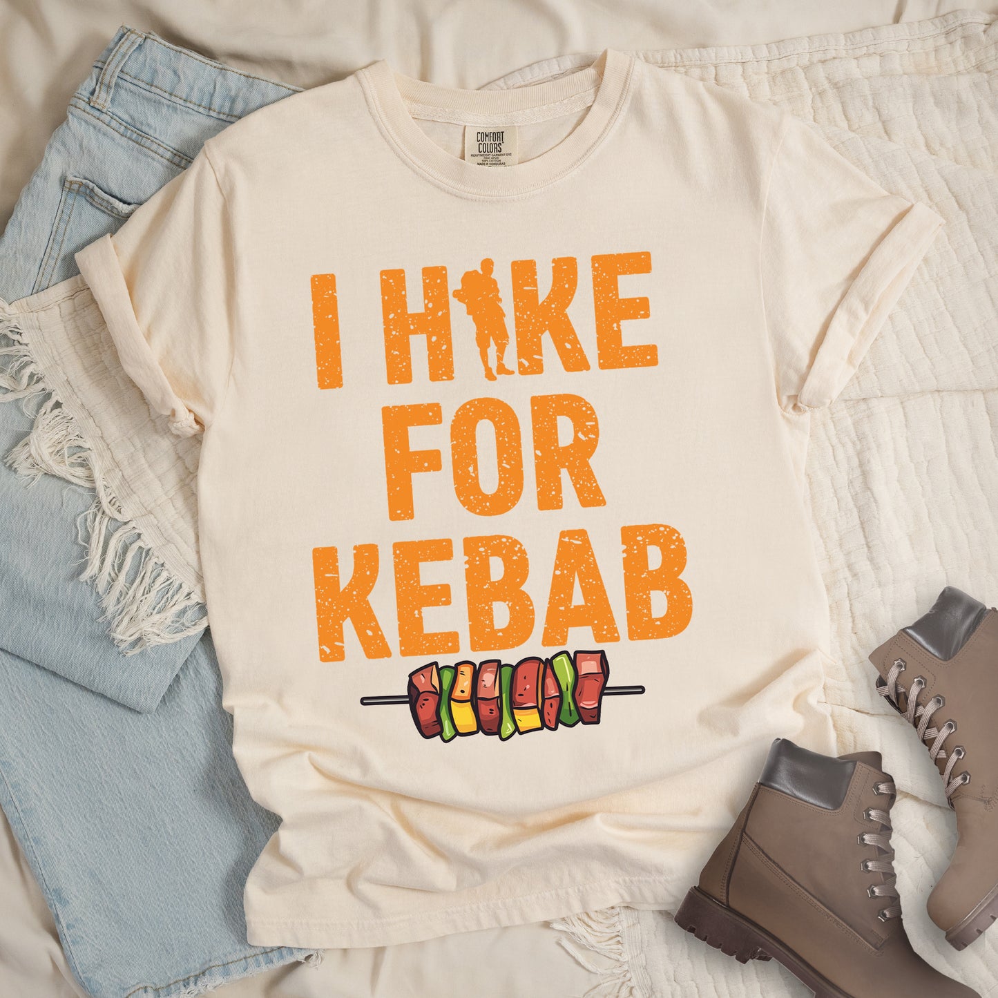 Ivory (Cream White) t-shirt with orange distressed text reading "I HIKE FOR KEBAB". "I HIKE" appears at the top with a hiker silhouette integrated into the letter "I", "FOR" in the middle, and "KEBAB" at the bottom. Below the text is a colorful illustration of a kebab skewer with meat and vegetables. Features a classic crew neck and casual rolled sleeves, styled alongside light wash denim and brown hiking boots.