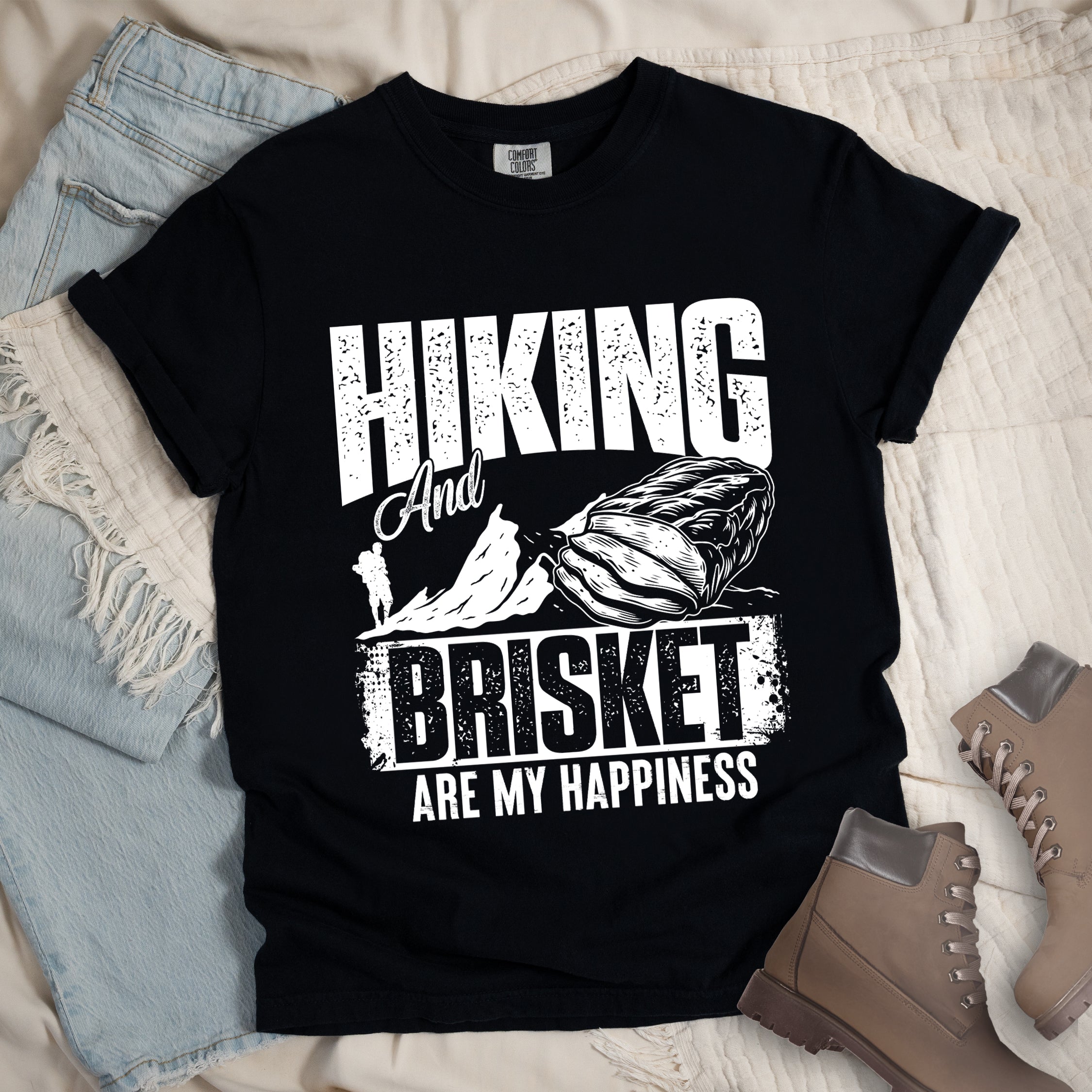 Black t-shirt with white distressed text reading "HIKING And BRISKET ARE MY HAPPINESS". "HIKING" appears in large letters at the top, "And" in script in the middle, "BRISKET" in bold distressed letters, and "ARE MY HAPPINESS" in smaller text at the bottom. Features a detailed white illustration of a brisket cut of meat on the right and mountain silhouettes with a small hiker figure on the left.