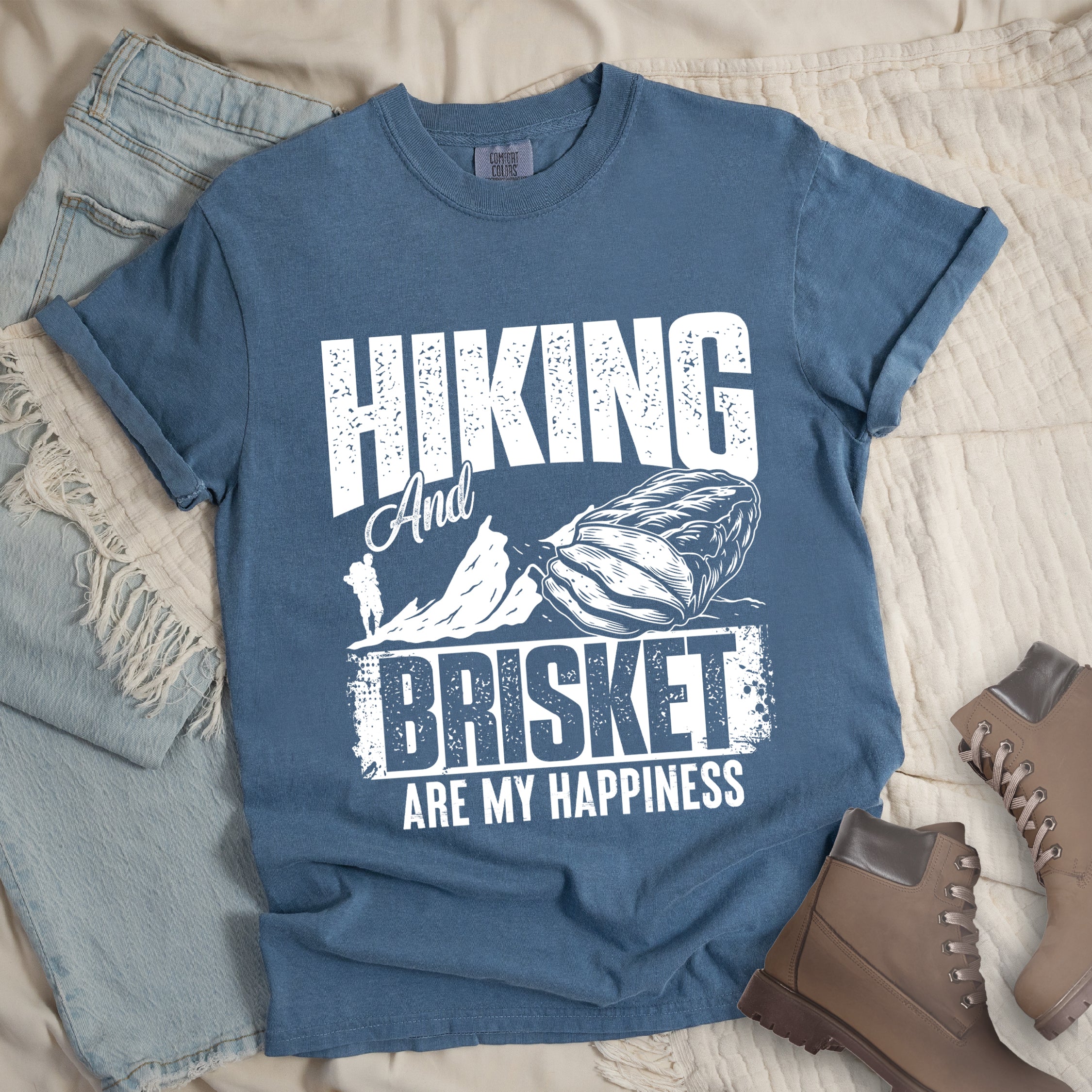 Blue Jean (Denim Blue) t-shirt with white distressed text reading "HIKING And BRISKET ARE MY HAPPINESS". "HIKING" appears in large letters at the top, "And" in script in the middle, "BRISKET" in bold distressed letters, and "ARE MY HAPPINESS" in smaller text at the bottom. Features a detailed white illustration of a brisket cut of meat on the right and mountain silhouettes with a small hiker figure on the left.