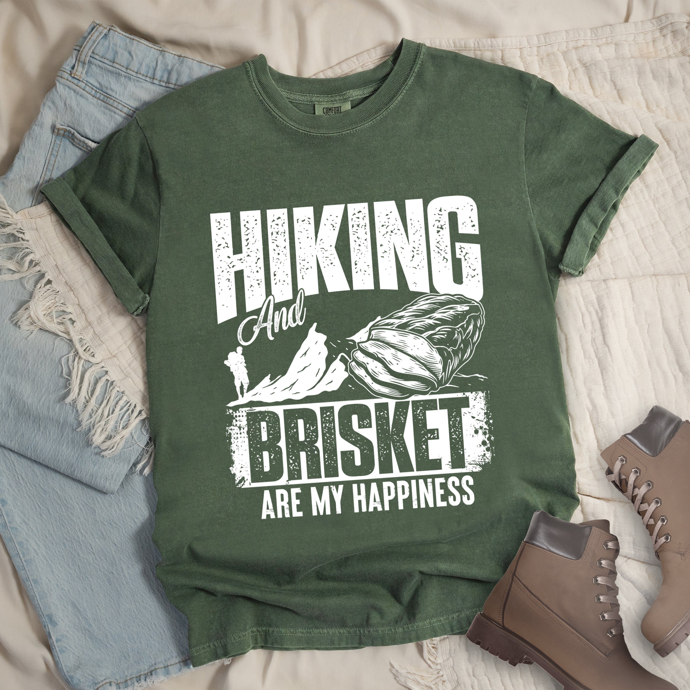 Moss (Dark Green) t-shirt with white distressed text reading "HIKING And BRISKET ARE MY HAPPINESS". "HIKING" appears in large letters at the top, "And" in script in the middle, "BRISKET" in bold distressed letters, and "ARE MY HAPPINESS" in smaller text at the bottom. Features a detailed white illustration of a brisket cut of meat on the right and mountain silhouettes with a small hiker figure on the left.