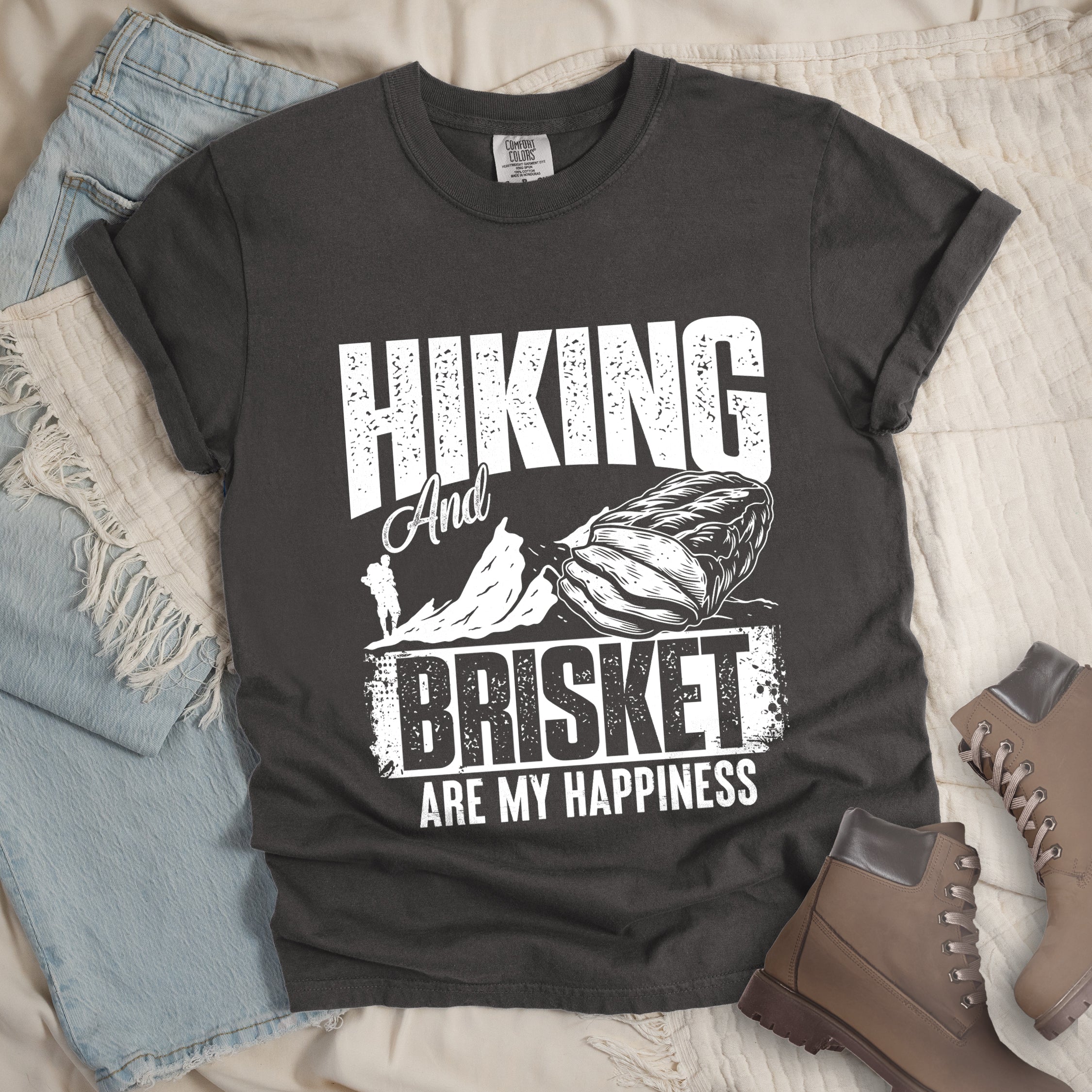 Pepper (Dark Gray) t-shirt with white distressed text reading "HIKING And BRISKET ARE MY HAPPINESS". "HIKING" appears in large letters at the top, "And" in script in the middle, "BRISKET" in bold distressed letters, and "ARE MY HAPPINESS" in smaller text at the bottom. Features a detailed white illustration of a brisket cut of meat on the right and mountain silhouettes with a small hiker figure on the left.