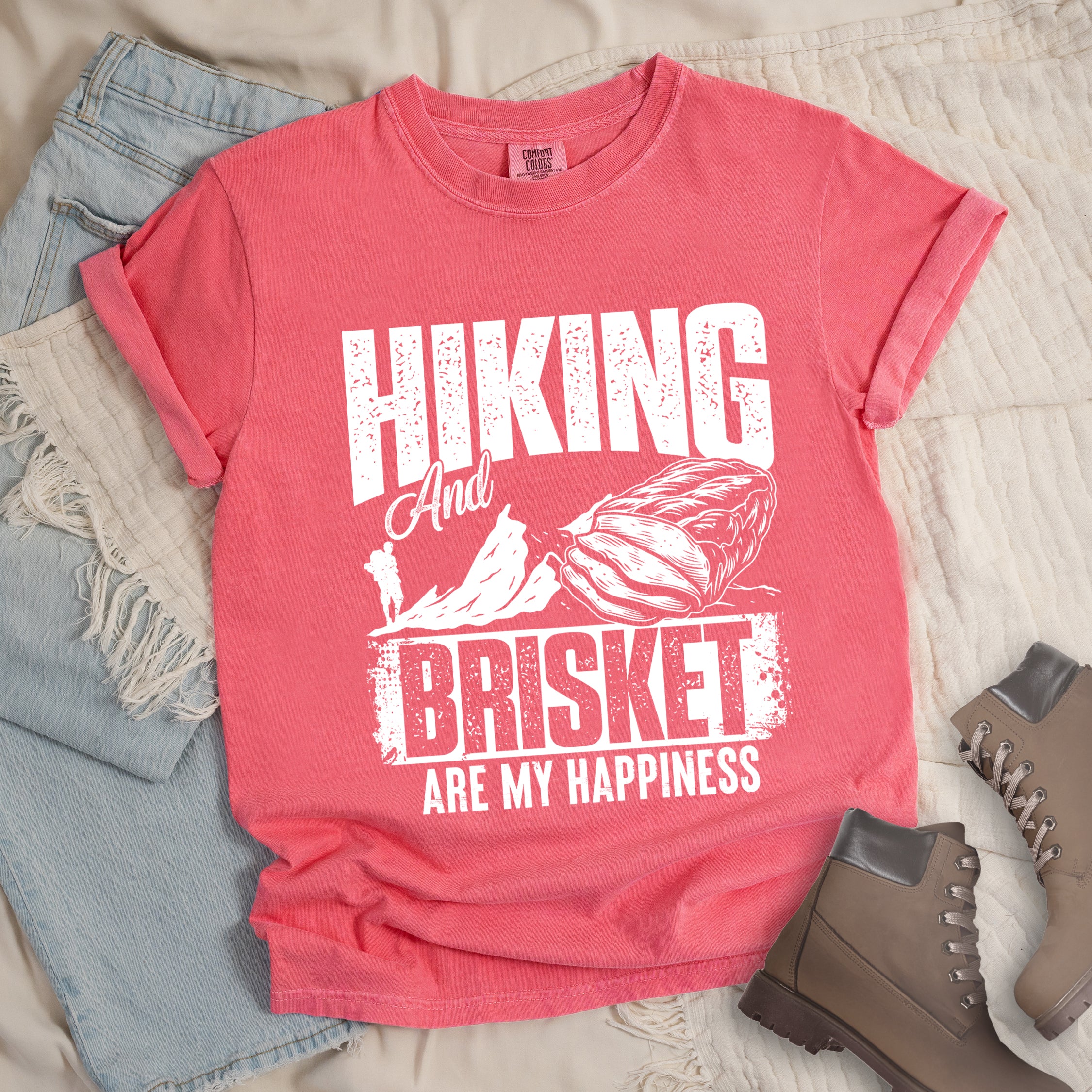 Watermelon (Light Coral) t-shirt with white distressed text reading "HIKING And BRISKET ARE MY HAPPINESS". "HIKING" appears in large letters at the top, "And" in script in the middle, "BRISKET" in bold distressed letters, and "ARE MY HAPPINESS" in smaller text at the bottom. Features a detailed white illustration of a brisket cut of meat on the right and mountain silhouettes with a small hiker figure on the left.