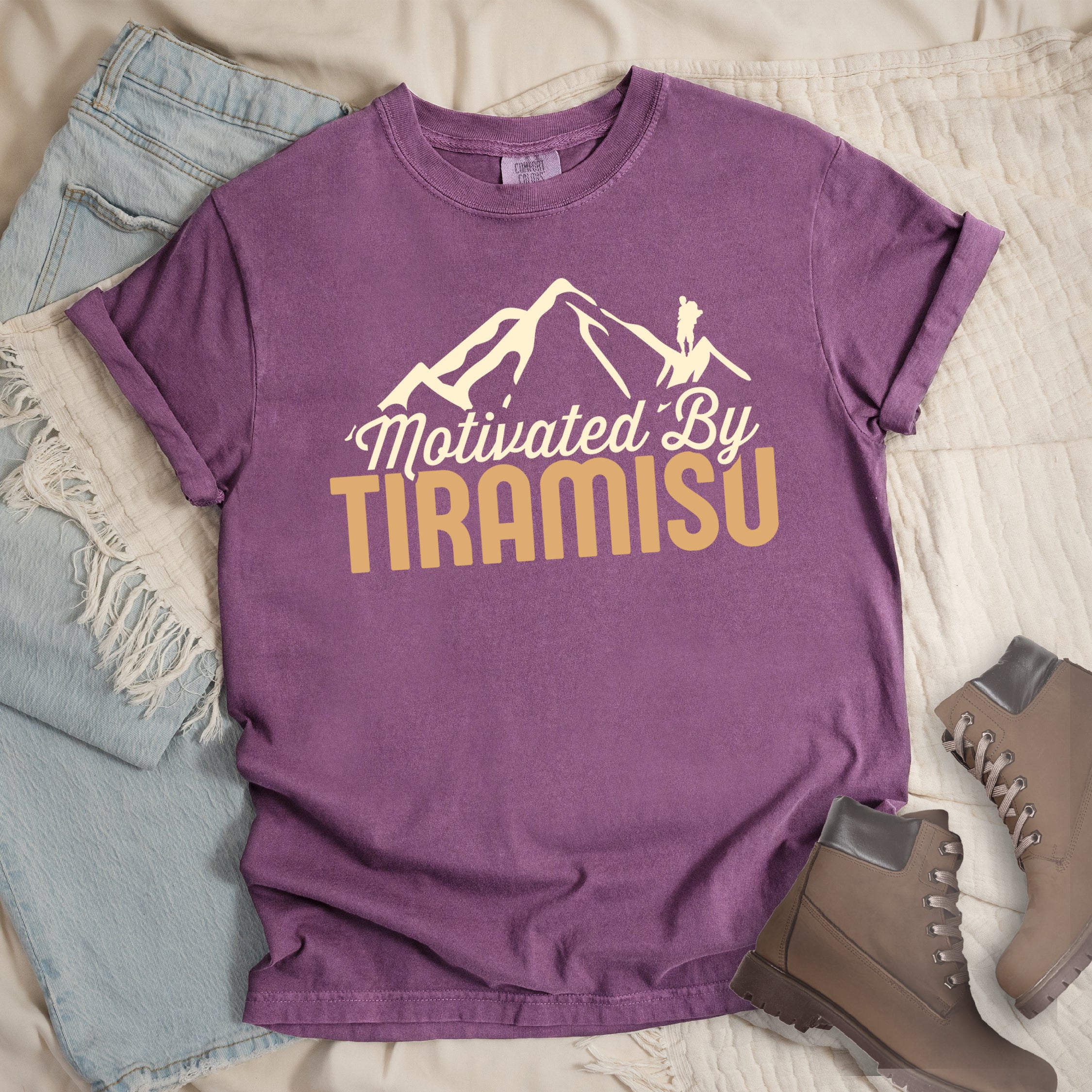 Berry (Dark Violet) t-shirt with cream and gold text reading "Motivated By TIRAMISU". "Motivated By" appears in cream script underneath a cream mountain silhouette with a small hiker figure on the peak, and "TIRAMISU" in large gold letters at the bottom. The shirt features a classic crew neck and casual rolled sleeves, styled alongside light wash denim and brown hiking boots. The design humorously connects the Italian coffee-flavored dessert as motivation for hiking and outdoor activities.