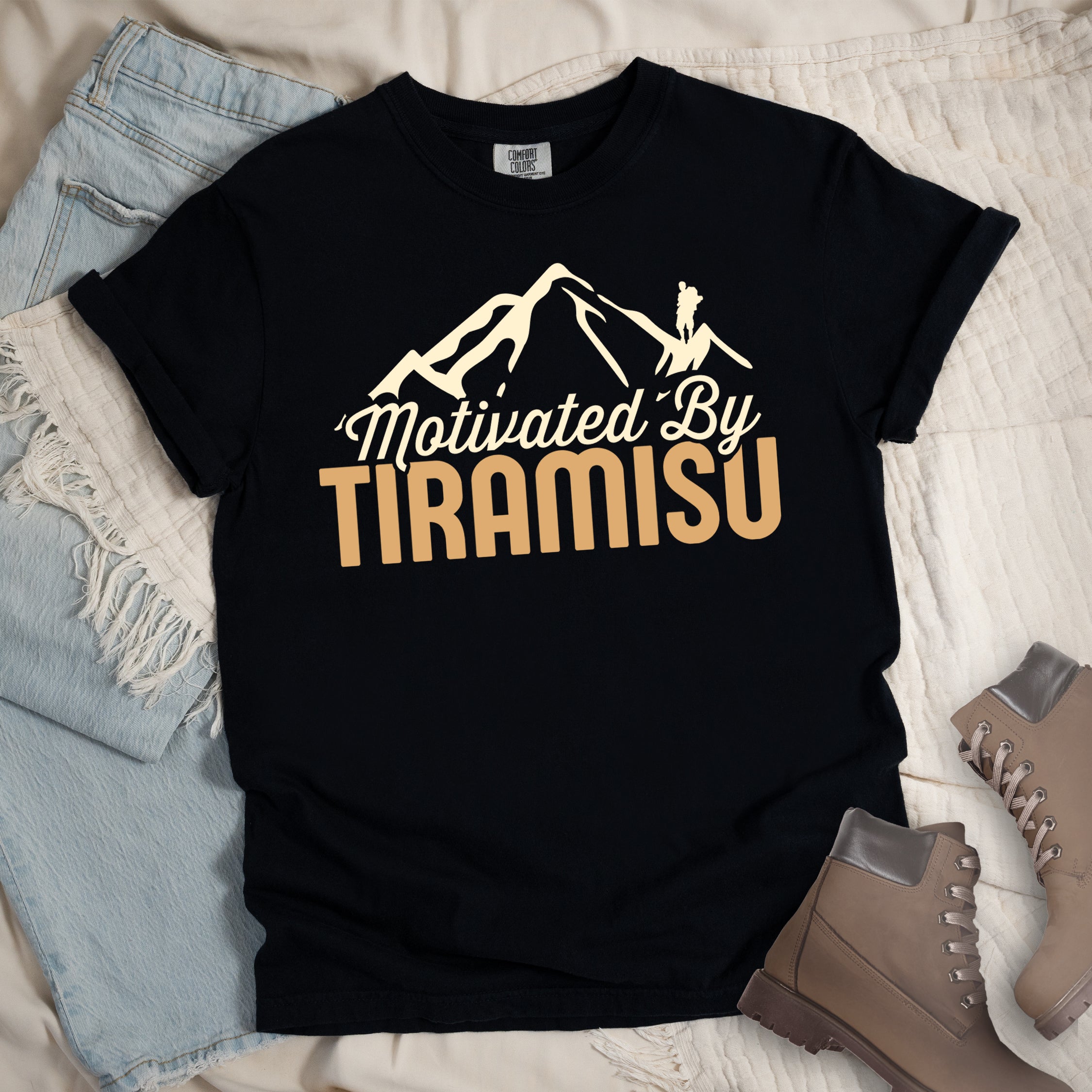 Black t-shirt with cream and gold text reading "Motivated By TIRAMISU". "Motivated By" appears in cream script underneath a cream mountain silhouette with a small hiker figure on the peak, and "TIRAMISU" in large gold letters at the bottom. The design humorously connects the Italian coffee-flavored dessert as motivation for hiking and outdoor activities. The shirt features a classic crew neck and casual rolled sleeves, styled alongside light wash denim and brown hiking boots.