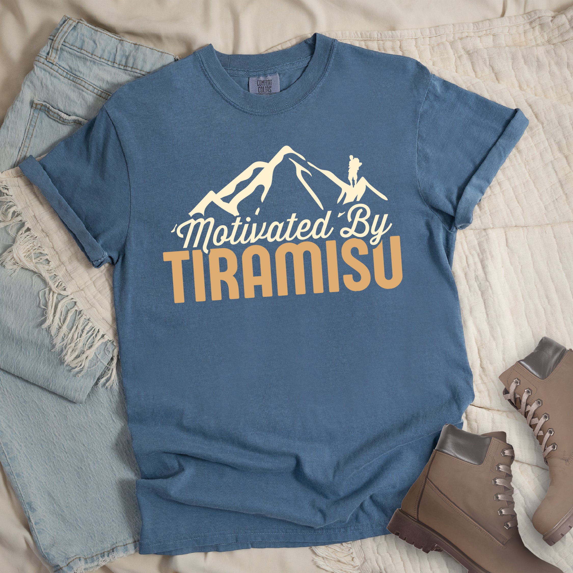 Blue Jean (Denim Blue) t-shirt with cream and gold text reading "Motivated By TIRAMISU". "Motivated By" appears in cream script underneath a cream mountain silhouette with a small hiker figure on the peak, and "TIRAMISU" in large gold letters at the bottom. The shirt features a classic crew neck and casual rolled sleeves, styled alongside light wash denim and brown hiking boots.The design humorously connects the Italian coffee-flavored dessert as motivation for hiking and outdoor activities.