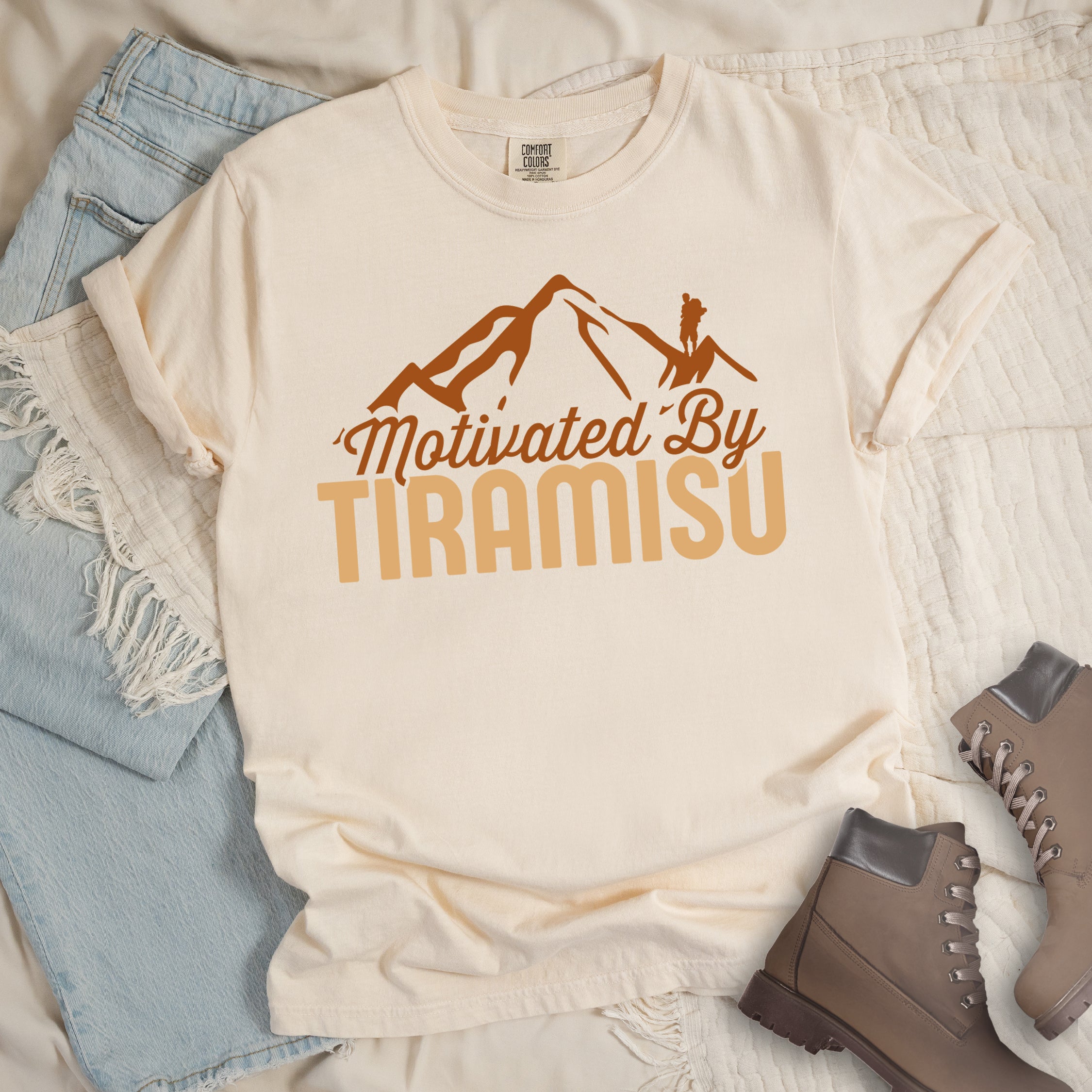 Ivory (Cream White) t-shirt with brown and gold text reading "Motivated By TIRAMISU". "Motivated By" appears in brown script underneath a brown mountain silhouette with a small hiker figure on the peak, and "TIRAMISU" in large gold letters at the bottom. The design humorously connects the Italian coffee-flavored dessert as motivation for hiking and outdoor activities. The shirt features a classic crew neck and casual rolled sleeves, styled alongside light wash denim and brown hiking boots.